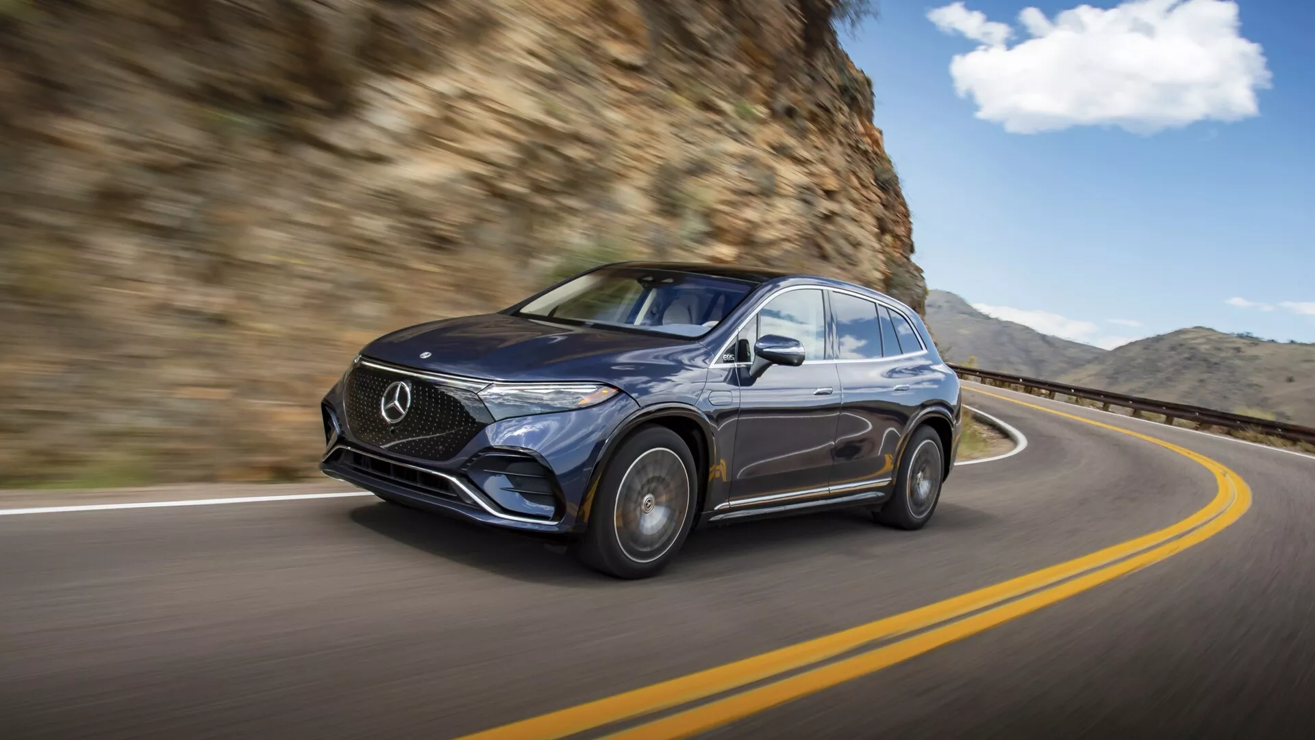 Obrázok tmavého Mercedesu EQS SUV 2022 v pohybe, zachytený z predného ľavého trojštvrťového pohľadu. Auto jazdí po zakrivenej ceste so skalnou stenou na pravej strane. Dôraz je kladený na luxusný dizajn s výraznou mriežkou s emblémom Mercedes, elegantnými svetlometmi, lesklým povrchom a čiastočne viditeľnými zliatinovými kolesami. Rozmazané pozadie zvyšuje pocit pohybu, podtrhujúc jazdné schopnosti a prémiový vzhľad vozidla v prirodzenom jazdnom prostredí.