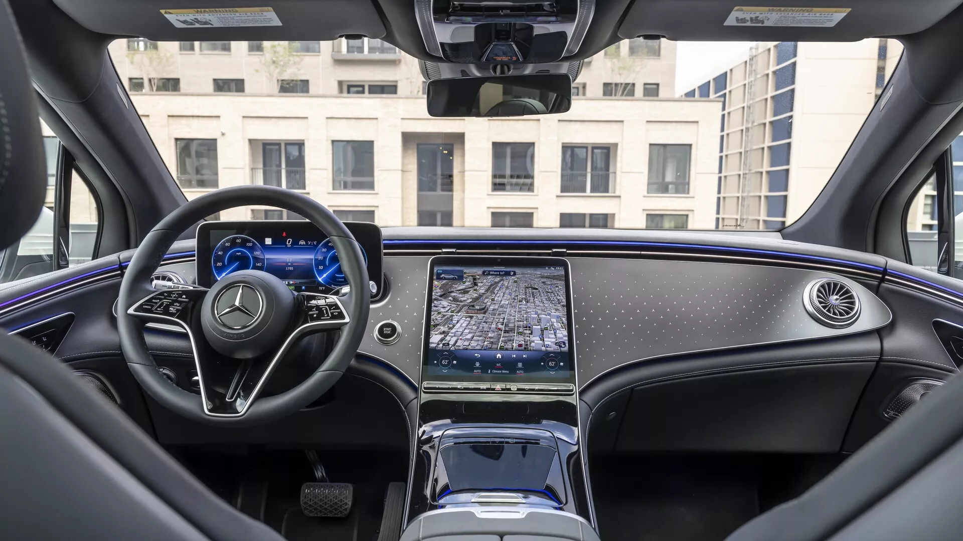 Interiérový pohľad na Mercedes EQS SUV 2022 z pohľadu zo zadného sedadla, zachytávajúci prístrojovú dosku, multifunkčný volant s plochým spodkom a kovovými akcentmi a stredovú konzolu s predĺženým dotykovým infotainment systémom. Digitálny prístrojový panel je osvetlený a minimalistický elektronický volič prevodových stupňov, úložné priestory a držiaky nápojov sú viditeľné. Luxusný interiér vozidla zahŕňa materiály vysokej kvality, leštené drevené obloženie a čiastočne viditeľnú panoramatickú strechu, ukazujúc moderný automobilový dizajn a pokročilé technologické funkcie.