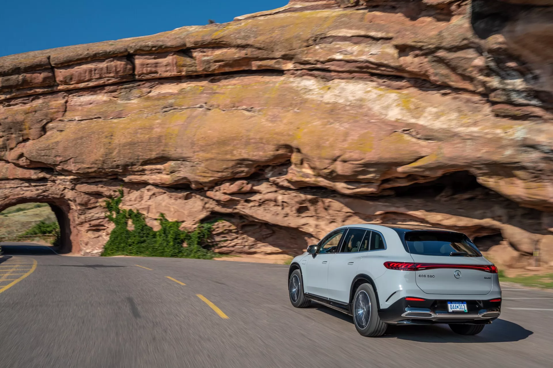 Biely Mercedes EQS-SUV 2022 jazdíci na zakrivenej dvojprúdovej ceste, fotografovaný zo zadného trojštvrťového pohľadu, ukazujúci zadné svetlá a bočný profil spolujazdca. Vlastnosti zahŕňajú výrazný zadný spojler, dvojitý výfukový systém, strieborné ozdobné lišty, chrómované akcenty a viaclúčové zliatinové kolesá. Rozmazanie pohybu na kolesách a pozadí zachytáva dynamickú jazdu SUV v malebnej horskej oblasti pod jasnou modrou oblohou.