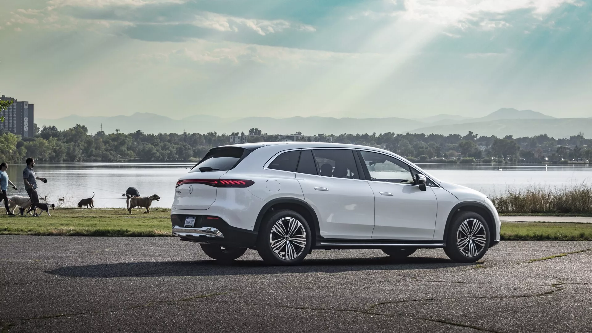 Profilový pohľad na luxusný Mercedes EQS SUV z roku 2022 fotografovaný na asfaltovej ceste. SUV má veľké viacpruhové zliatinové kolesá, elegantnú líniu okien zužujúcu sa smerom dozadu s tónovanými oknami, výrazné aerodynamické línie karosérie, pokročilé bočné zrkadlá, zvažujúcu sa strechu s nosičom a ovíjajúce sa LED zadné svetlá. Vozidlo je zasadené do teplého slnečného svetla, čo zdôrazňuje jeho moderný dizajn v pokojnom prostredí s telom vody, stromami a kráčajúcimi ľuďmi.