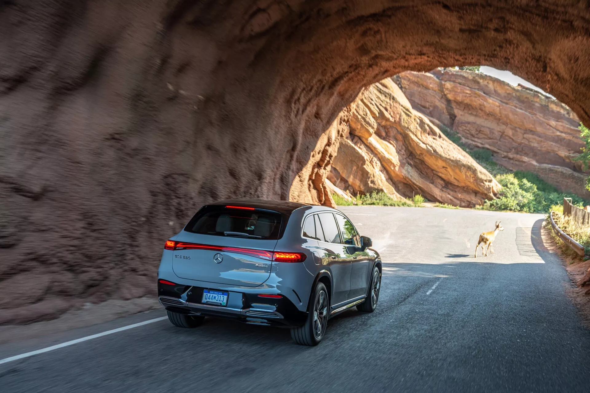 Zadný a uhlový pohľad na elegantné, moderné SUV prechádzajúce cez skalný tunel za slnečného dňa. Kľúčové prvky zahŕňajú horizontálne obopínajúce zadné svetlá, športový spojler, žraločiu anténu, reflexné okenné lišty, dvojité výfukové potrubia, päťlúčové zliatinové kolesá a centrálne umiestnenú ŠPZ pod tónovaným zadným oknom. Vozidlo vykazuje pokročilú osvetľovaciu technológiu a pravdepodobne zahŕňa funkcie ako cúvacia kamera a parkovacie senzory. Fotografia zachytáva pohyb a pocit outdoorového dobrodružstva s jeleňom v pozadí.