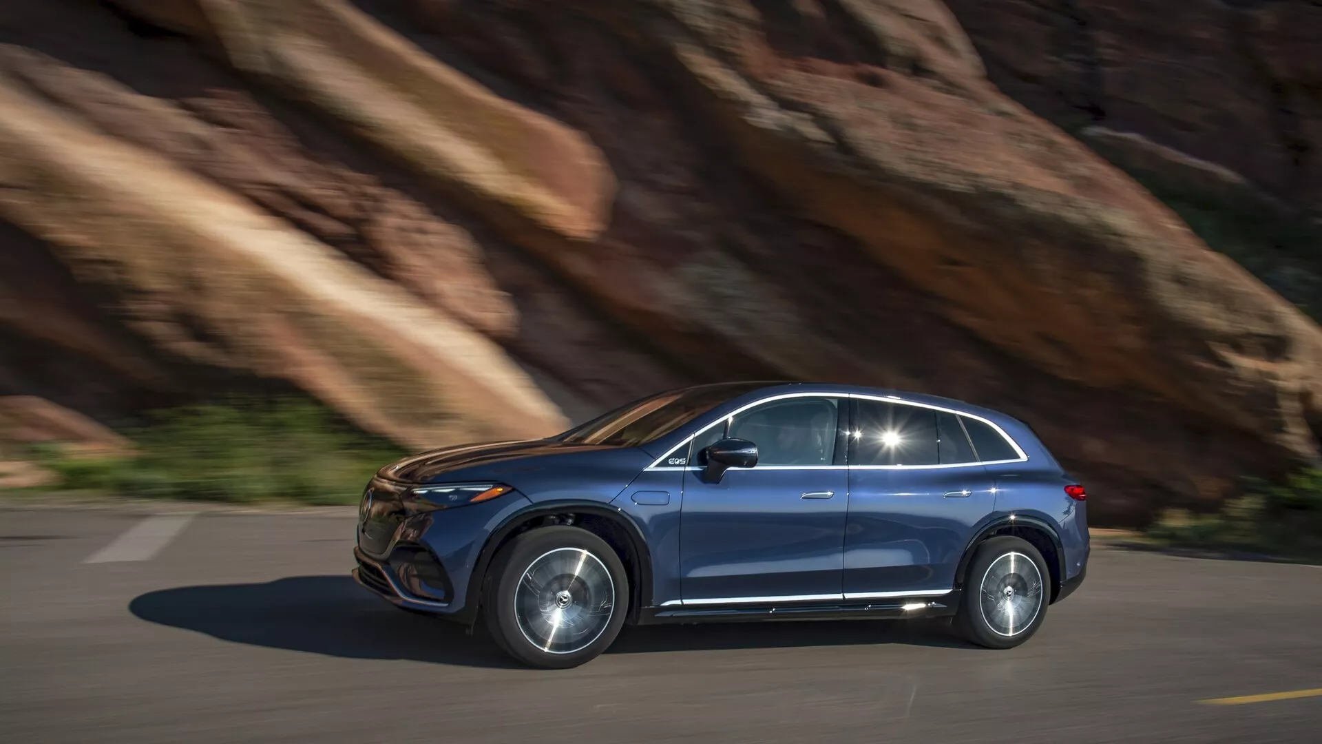 Mercedes EQS-SUV 2022 v pohybe, bočný profil. Jazda zľava doprava po asfaltovej ceste s pozadím skalného útesu, naznačujúcim horský terén. Pravý bočný pohľad zobrazuje moderný dizajn: viditeľné predné a zadné dvere pre cestujúcich, dobre definované bočné okná s rámom, zliatinové kolesá a výrazné bočné zrkadlo. Kovový lak, pravdepodobne LED alebo HID svetlá, zapustené kľučky dverí. Možné pokročilé technologické funkcie: bezpečnostné systémy, infotainment, elektrický/hybridný motor. Menšie rozmazanie pohybu naznačuje dynamický výkon, denné svetlo pri prirodzenom osvetlení.