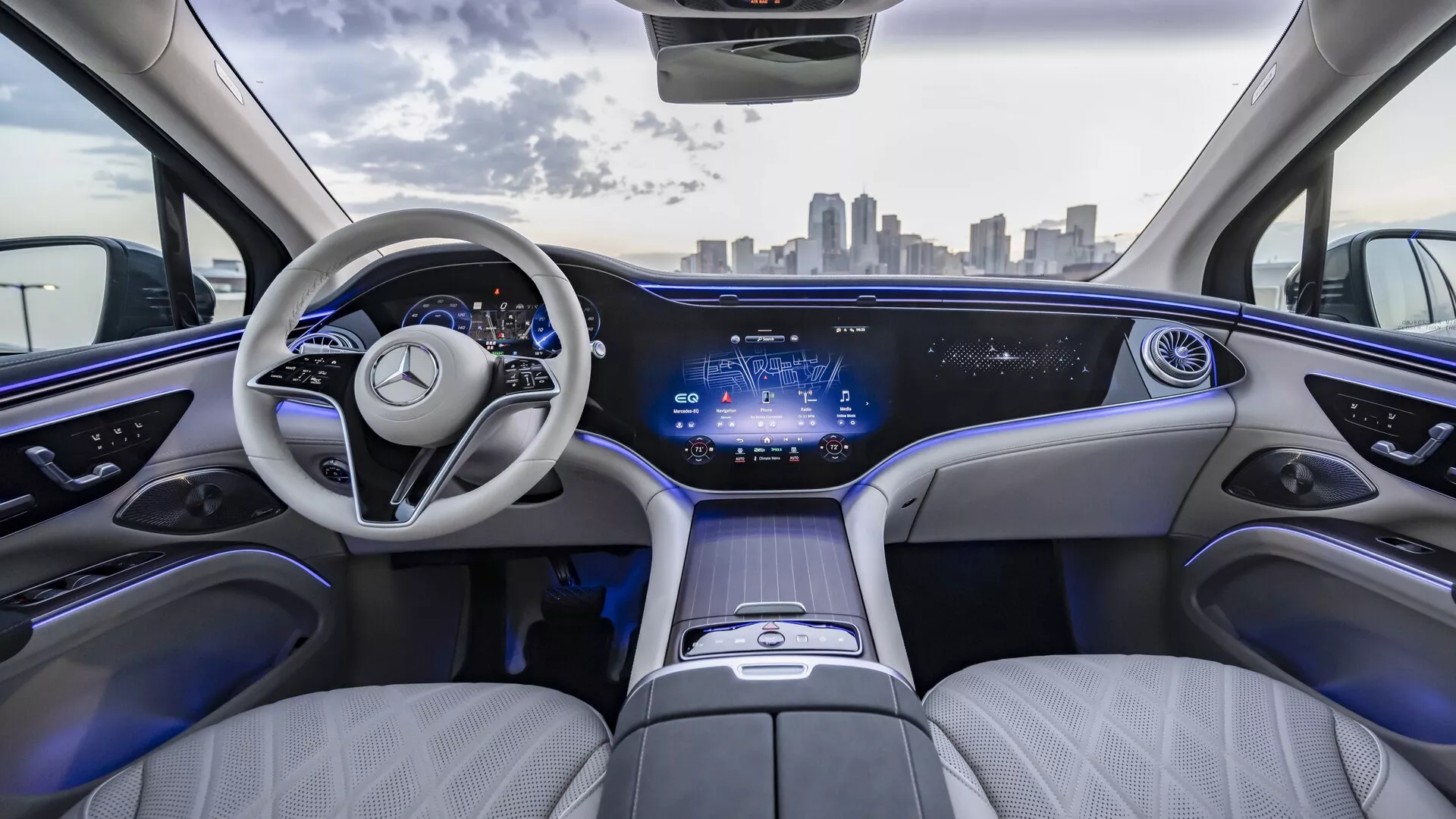 Pohľad na interiér Mercedesu EQS SUV 2022 zo zadných sedadiel, s luxusným svetlým koženým čalúnením, elegantnou palubnou doskou s kontinuálnym digitálnym displejom, moderným dvojramenným volantom s emblémom Mercedes-Benz, priestranným a pohodlným usporiadaním sedadiel, ambientným osvetlením, stredovou konzolou s dotykovým ovládaním a panoramatickým strešným oknom. Mestská panoráma viditeľná cez čelné sklo.