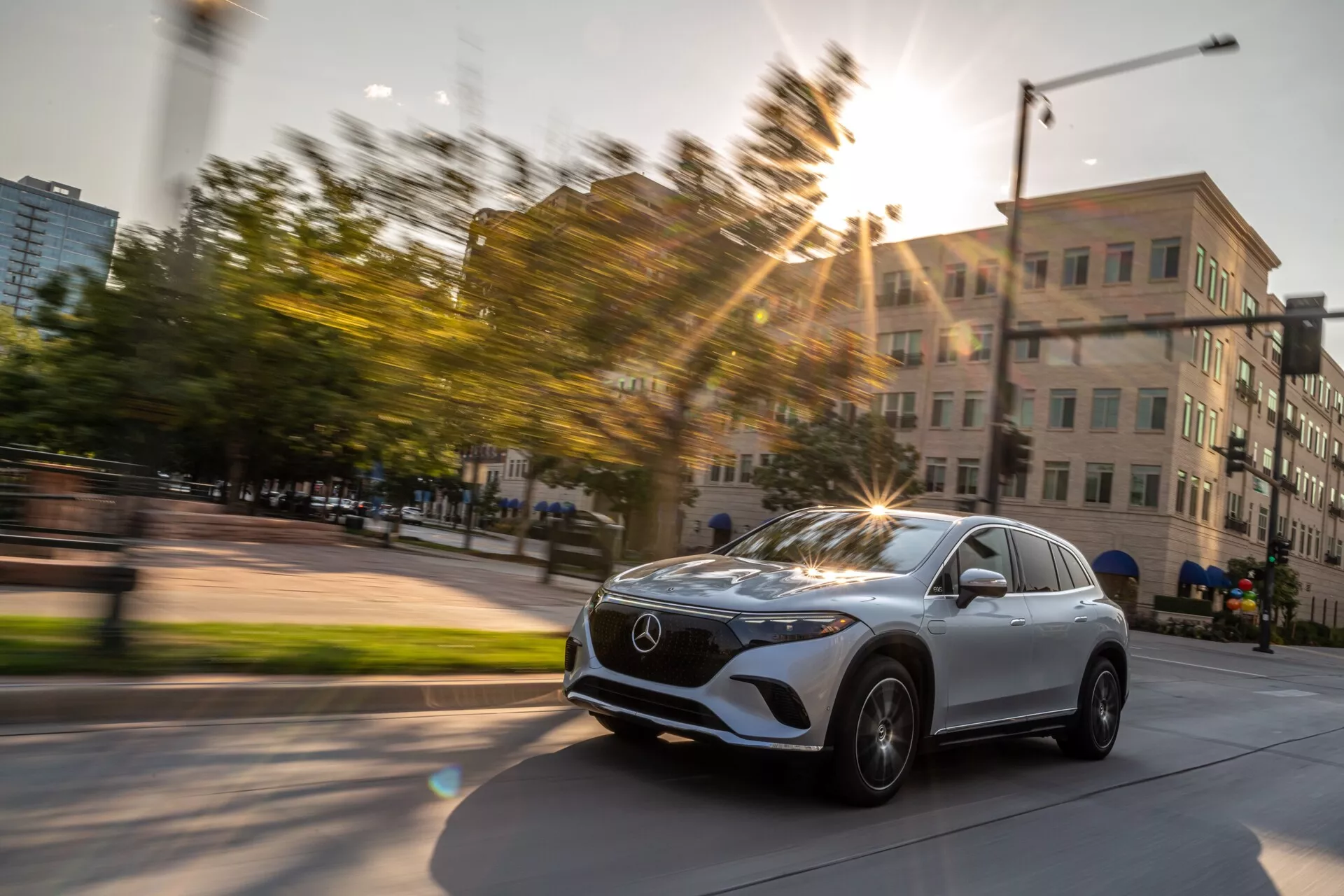 Dynamický obrázok 2022 Mercedes EQS SUV zachytený v pohybe z mierne šikmého predného trojštvrtinového pohľadu. Fotografia zvýrazňuje prednú mriežku vozidla s emblémom Mercedes, elegantné LED svetlomety, koleso a blatník na vodičovej strane a časť čelného skla a vodičových dverí. Kolesá auta sú otočené doprava, čo naznačuje zákrutu alebo zmenu smeru. Slnečné svetlo vytvára efekt svetelného závoja, ktorý dodáva energiu do fotografie, zhotovenej v mestskom prostredí s budovami a zeleňou počas zlatej hodiny. Pohybové rozmazanie v pozadí zdôrazňuje rýchlosť a pohyb.