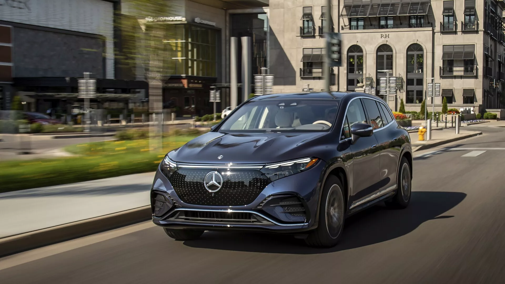 Dynamický záber na úrovni ulice, zachytávajúci SUV Mercedes EQS z roku 2022 v pohybe, snímaný z nízkej perspektívy z troch štvrtín spredu na strane vodiča. Luxusné vozidlo zdôrazňuje svoju výraznú prednú mriežku s veľkým emblémom, elegantné LED svetlomety s denným svietením a aerodynamický dizajn. Kolesá z ľahkých zliatin a bočné zrkadlá vo farbe karosérie so zabudovanými smerovkami zdôrazňujú luxus. Mestsé pozadie s rozmazaným pohybom zdôrazňuje pokročilé technologické prvky vozidla a jeho prítomnosť na ceste.