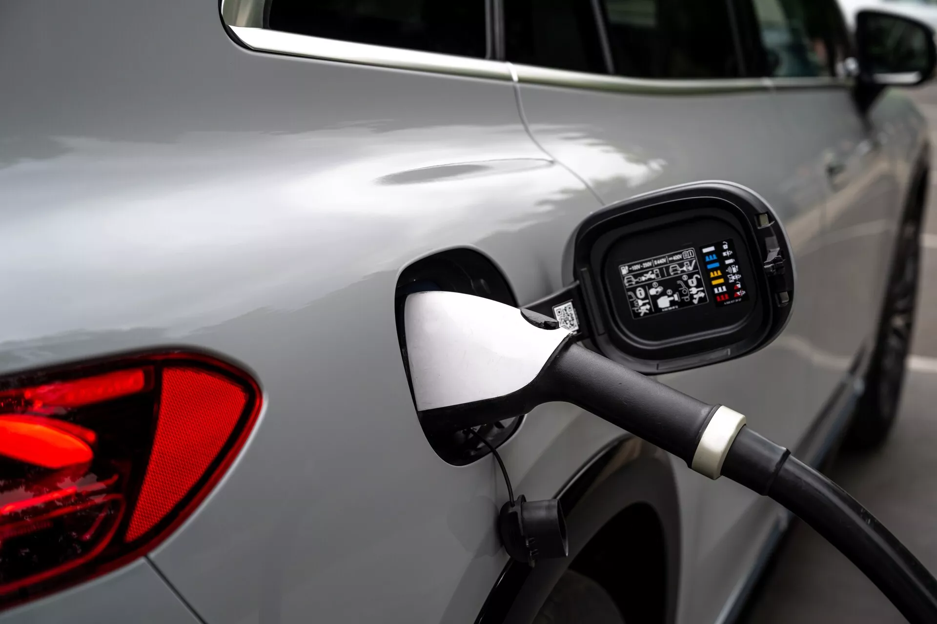 Detailný záber na oblasť nabíjacieho portu na boku strieborného Mercedes EQS-SUV 2022, zameraný na pripojený nabíjací kábel, otvorený port s digitálnym displejom indikujúcim stav nabíjania, s mierne rozmazaným pozadím zobrazujúcim časť zadného svetla a odraz iného vozidla.