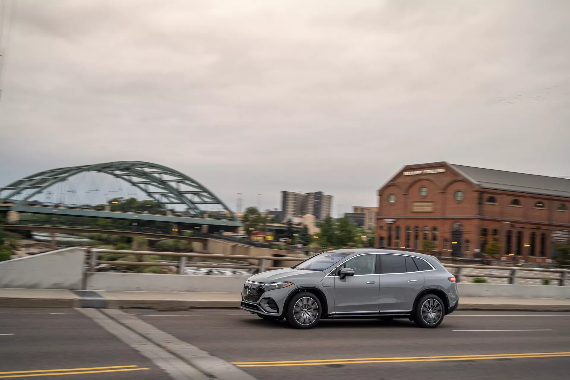 Bočný profilový pohľad na moderný Mercedes EQS-SUV 2022, ktorý jazdí po mestskej ulici, ukazujúci elegantný dizajn, päť dverí, hliníkové kolesá a LED svetlomety. Zachytené z mierne zadného uhla s rozmazaným pozadím.