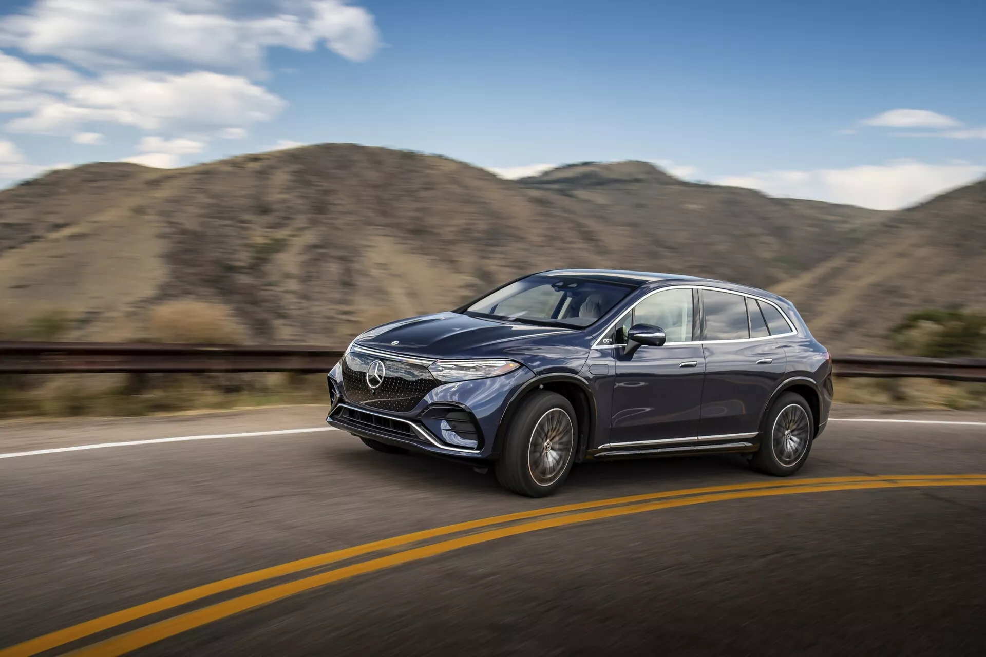 Záber v pohybe moderného Mercedesa EQS-SUV 2022 prechádzajúceho po hladkej asfaltovej ceste so žltou deliacou čiarou pod jasnou oblohou. Auto je vidieť z mierneho predného bočného uhla, zdôrazňujúc vodičovu stranu, vrátane prednej mriežky, dizajnu svetlometov, dverí na strane vodiča, bočného zrkadla a časti čelného skla. Vozidlo je zaostrené s rozmazaným pozadím s kopcami, čo naznačuje pohyb. Viditeľné sú dizajnové prvky ako zliatinové kolesá, tvarované bočné panely, výrazné štýlové línie a potenciálna technológia ADAS.