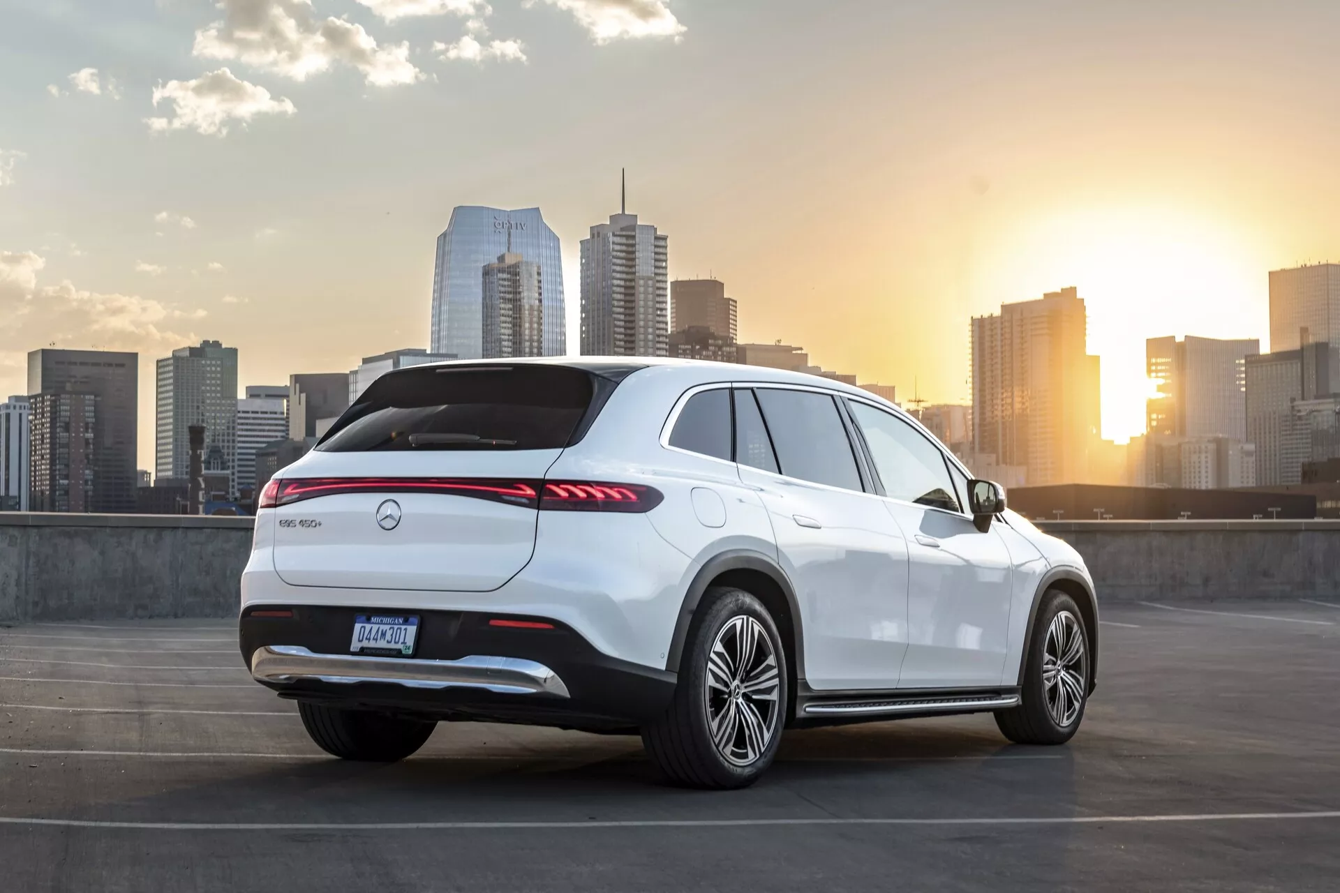 Obrázok bieleho Mercedes EQS-SUV 2022 zaparkovaného na najvyššej úrovni parkovacej štruktúry. Vozidlo je vidieť z trojštvrťovej zadnej perspektívy, ukazujúce zadnú, bočnú stranu a časť strešnej línie. Obsahuje predĺžené zadné LED svetlá, emblém výrobcu uprostred zadného veka, zadný difúzor, zadný spojler, zliatinové kolesá a vysokú líniu pásu s klesajúcou športovou strechou. Scéna zahŕňa panorámu mesta a západ slnka v pozadí, zvýrazňujúc mestský luxus a moderný životný štýl.