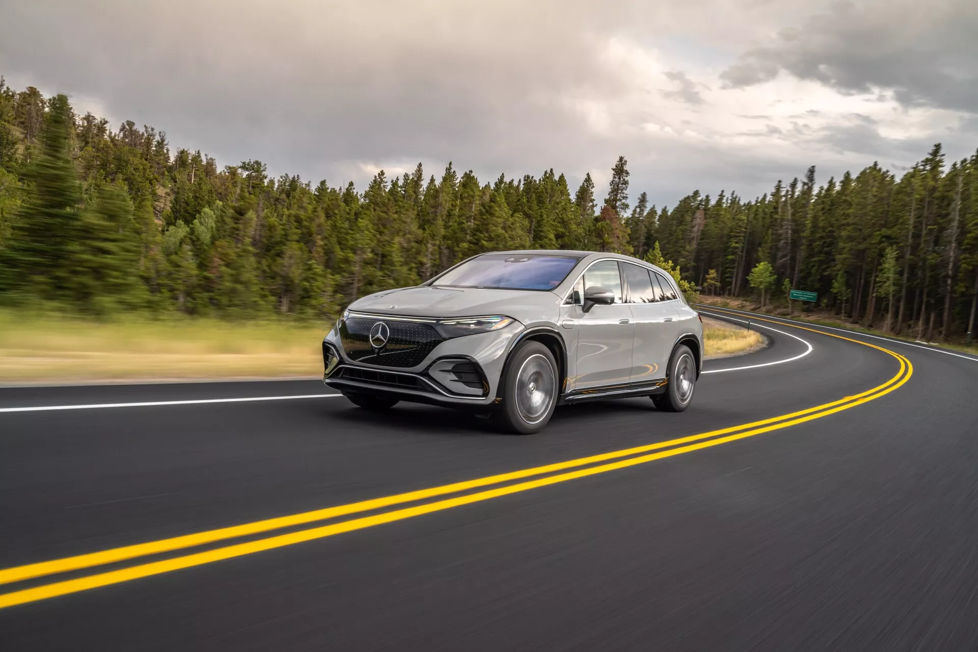 Tento obrázok zachytáva Mercedes-Benz EQS SUV 2022 v pohybe z trojštvrťového predného uhla s viditeľným predným dizajnom a bočným profilom. Vozidlo je umiestnené vľavo, jazdí po dobre udržiavanej asfaltovej ceste s dvojitými žltými čiarami a pravotočivou zákrutou. Rozmazanie kolies a mierne rozmazanie spôsobené pomalšou rýchlosťou uzávierky zdôrazňujú rýchlosť a dynamický pohyb. Zatažené svetelné podmienky vytvárajú mäkké tiene a rovnomerné osvetlenie vozidla. Sú viditeľné technologické prvky, ako sú LED svetlá, denné svietenie, veľká predná maska s emblémom a aerodynamické bočné zrkadlá s možnými senzormi. Strieborný alebo svetlosivý lak kontrastuje s lesklou čiernou spodnou časťou masky, lemami blatníkov a okennými lištami, čo zdôrazňuje elegantné línie, zakrivené blatníky a strechu podobnú kupé. Vhodné pre automobilové, technologické, fotografické a luxusné vozidlá.