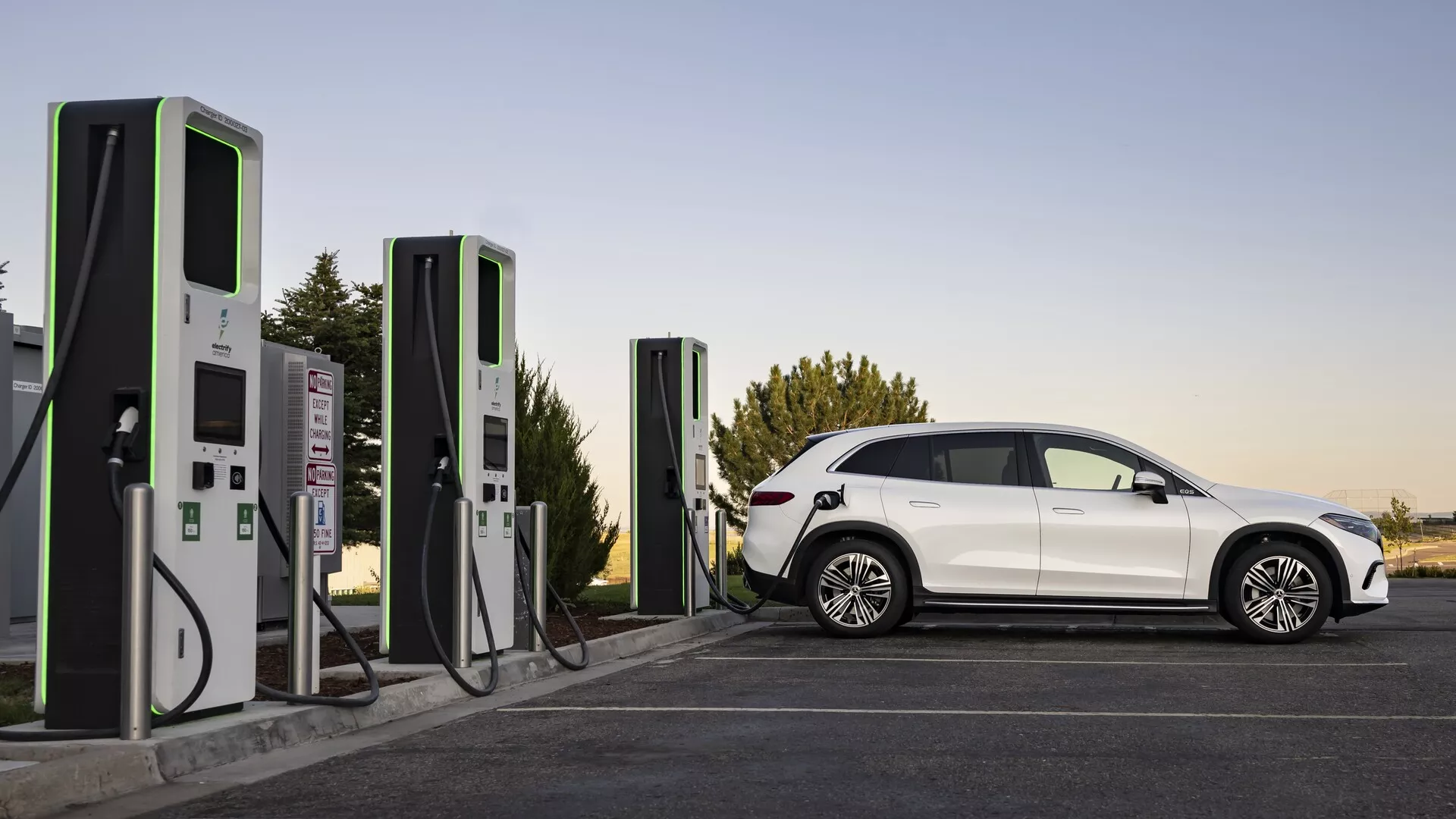 Elegantné a moderné elektrické vozidlo (EV), Mercedes EQS-SUV 2022, zobrazené pri nabíjacej stanici za mäkkých svetelných podmienok, buď neskoro popoludní alebo skoro ráno. Kompletný bočný profil odhaľuje aerodynamické línie, výrazné kolesá, zatmavené okná a dobre definovaný priestor pre pasažierov. Vozidlo je pripojené k modernej nabíjacej stanici, čím zdôrazňuje zameranie na udržateľnú dopravu a infraštruktúru pre elektromobily.
