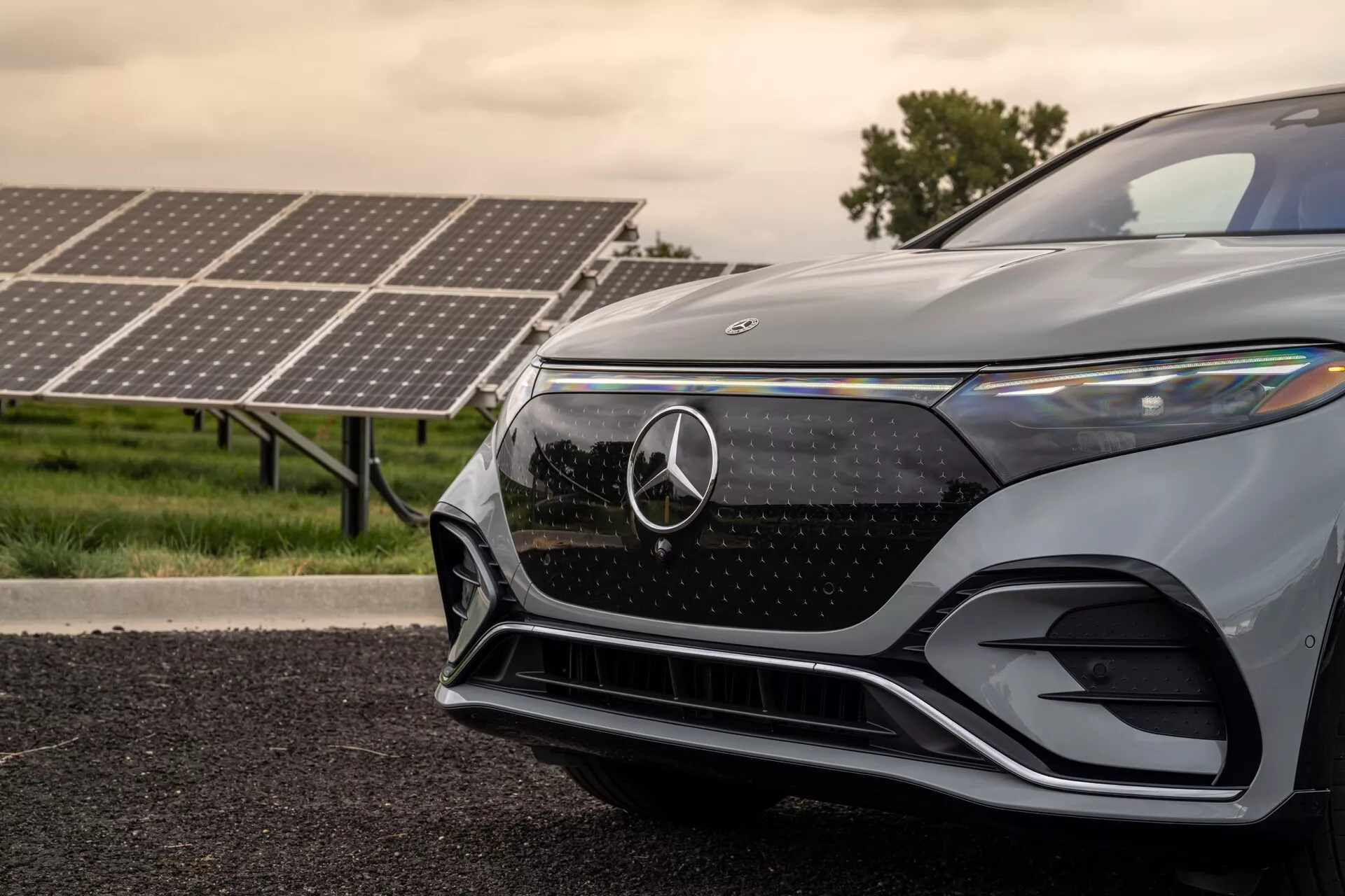 Detailný pohľad na predný ľavý štvrtok moderného Mercedes-Benz EQS SUV 2022 z nízkeho uhla. Obrázok zvýrazňuje mriežku so znakom Mercedes-Benz, štýlový uhlový LED svetlomet, aerodynamický predný nárazník a metalický lak. Solárne panely v pozadí naznačujú elektrickú alebo hybridnú povahu vozidla, zdôrazňujúce jeho pokrokové a ekologické vlastnosti.