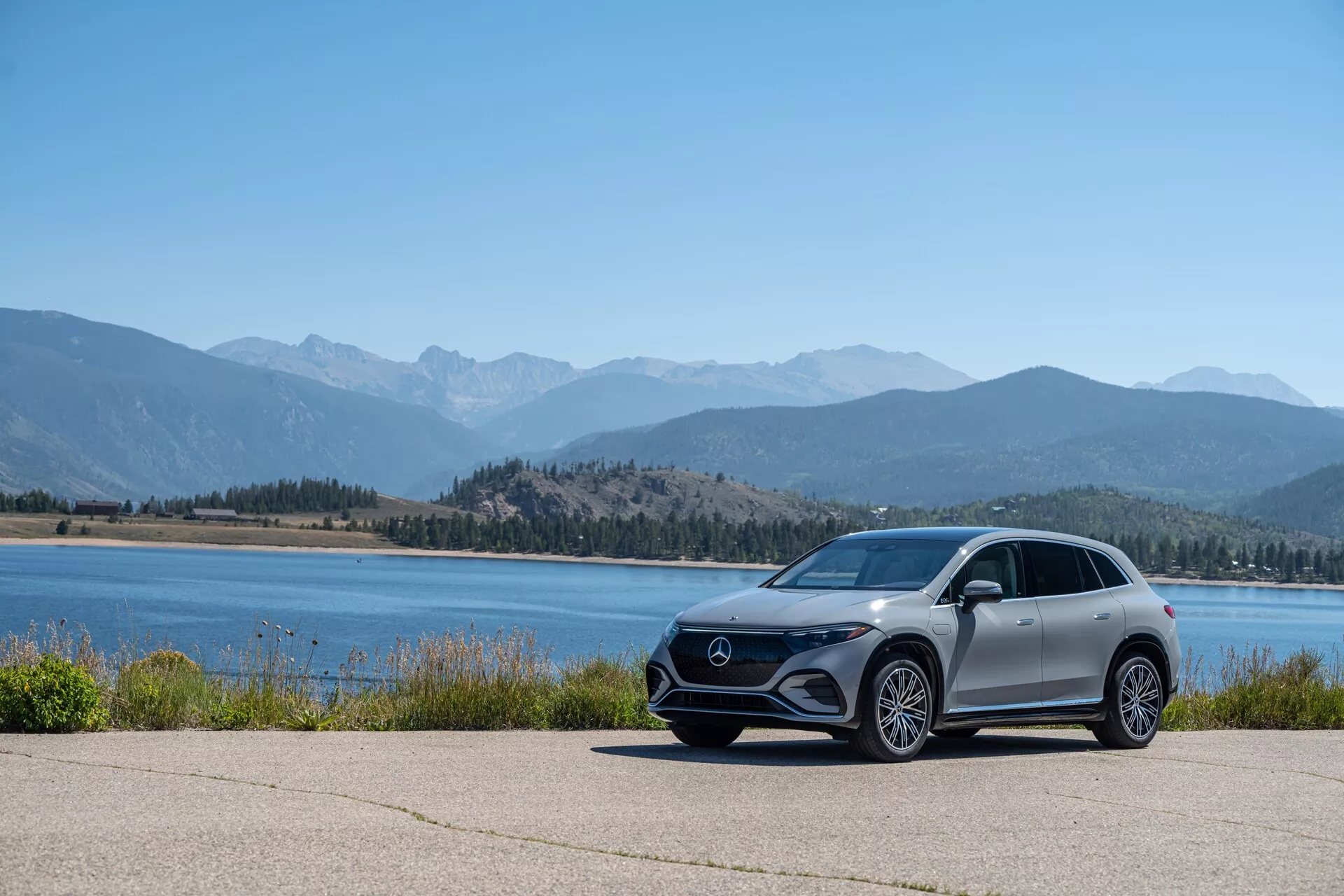 Obrázok zobrazuje moderné vozidlo Mercedes EQS SUV 2022 zaparkované na vydláždenej ploche s malebným pozadím vody a horských pásov, fotografované z nízkeho uhla na zdôraznenie jeho postoja a dizajnových línií. Auto je zobrazené z bočného profilu s viditeľnou prednou mriežkou, uhlovými LED svetlami, veľkými zliatinovými kolesami, aerodynamickými bočnými zrkadlami a metalickým lakovaním.