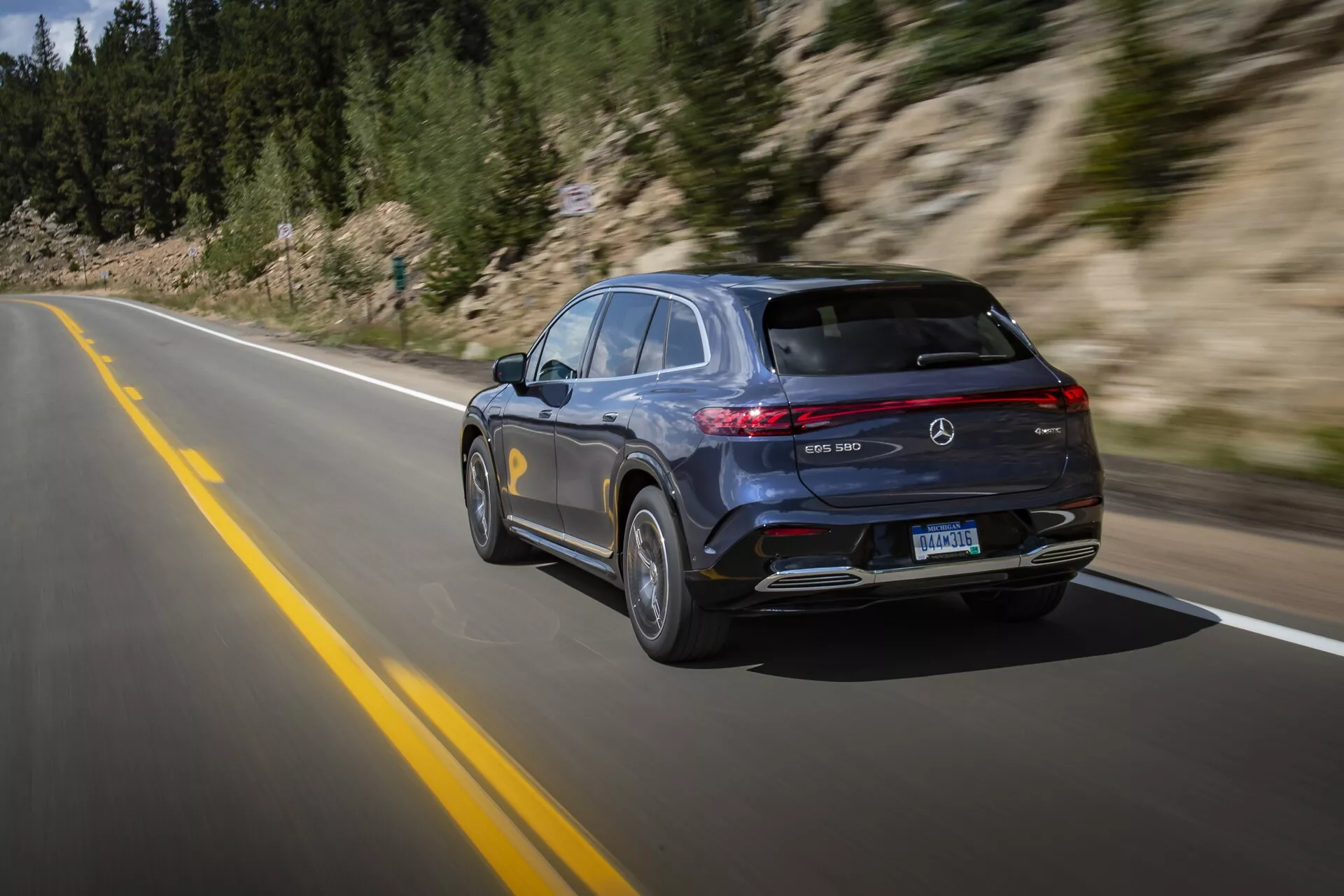 Moderný, luxusný Mercedes-Benz EQS SUV 2022 zachytený v pohybe na asfaltovej ceste, so štýlovým dizajnom s tmavým lakom, horizontálnymi LED zadnými svetlami, strešným spoilerom, dvojitými výfukmi a viacramennými zliatinovými kolesami. Technika paningu vo vysokej rýchlosti zvýrazňuje pohyb vozidla prírodným, horským prostredím.