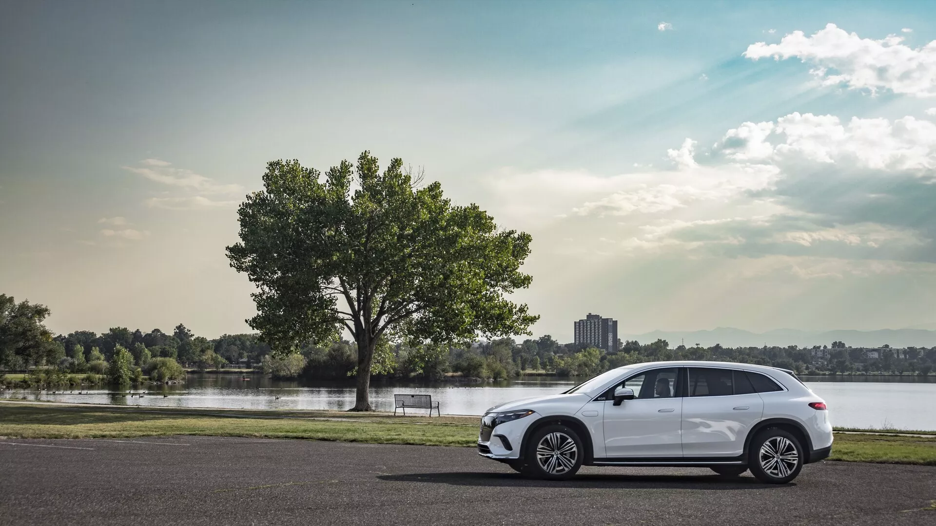Bočný profilný pohľad na moderný Mercedes EQS SUV 2022 zaparkovaný na asfalte, s viaclúčovými zliatinovými kolesami, elegantnými aerodynamickými líniami, štyrmi dverami, zatmavenými oknami s integrovaným smerovkou v bočnom zrkadle, športová línia strechy s strešnými nosičmi a čiastočne viditeľnými zadnými LED svetlami, zasadený do pokojného scenického pozadia s jazerom alebo riekou, stromom, trávou a čiastočne zamračenou oblohou.