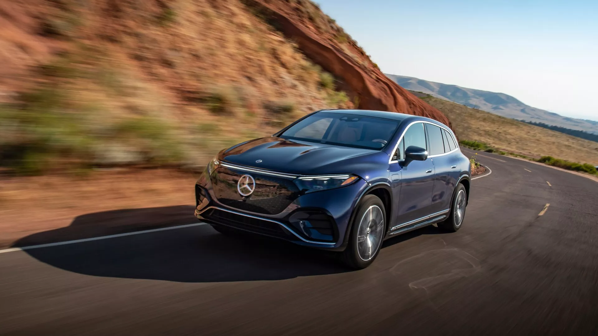 Dynamický záber Mercedesu EQS-SUV 2022 zachytený z trojštvrťového ľavého predného uhla, ktorý ukazuje vozidlo v pohybe. Medzi hlavné vlastnosti patrí elegantná ľavá strana auta, predná kapota, svetlomety s pokročilou LED technológiou, mriežka, predný nárazník a zliatinové kolesá. Aerodynamický dizajn karosérie naznačuje luxus a pokročilú techniku, zdôrazňuje výkon a zážitok z jazdy. Obraz vyžaruje pocit slobody a výkonu na čistých cestách obkolesených prírodou.