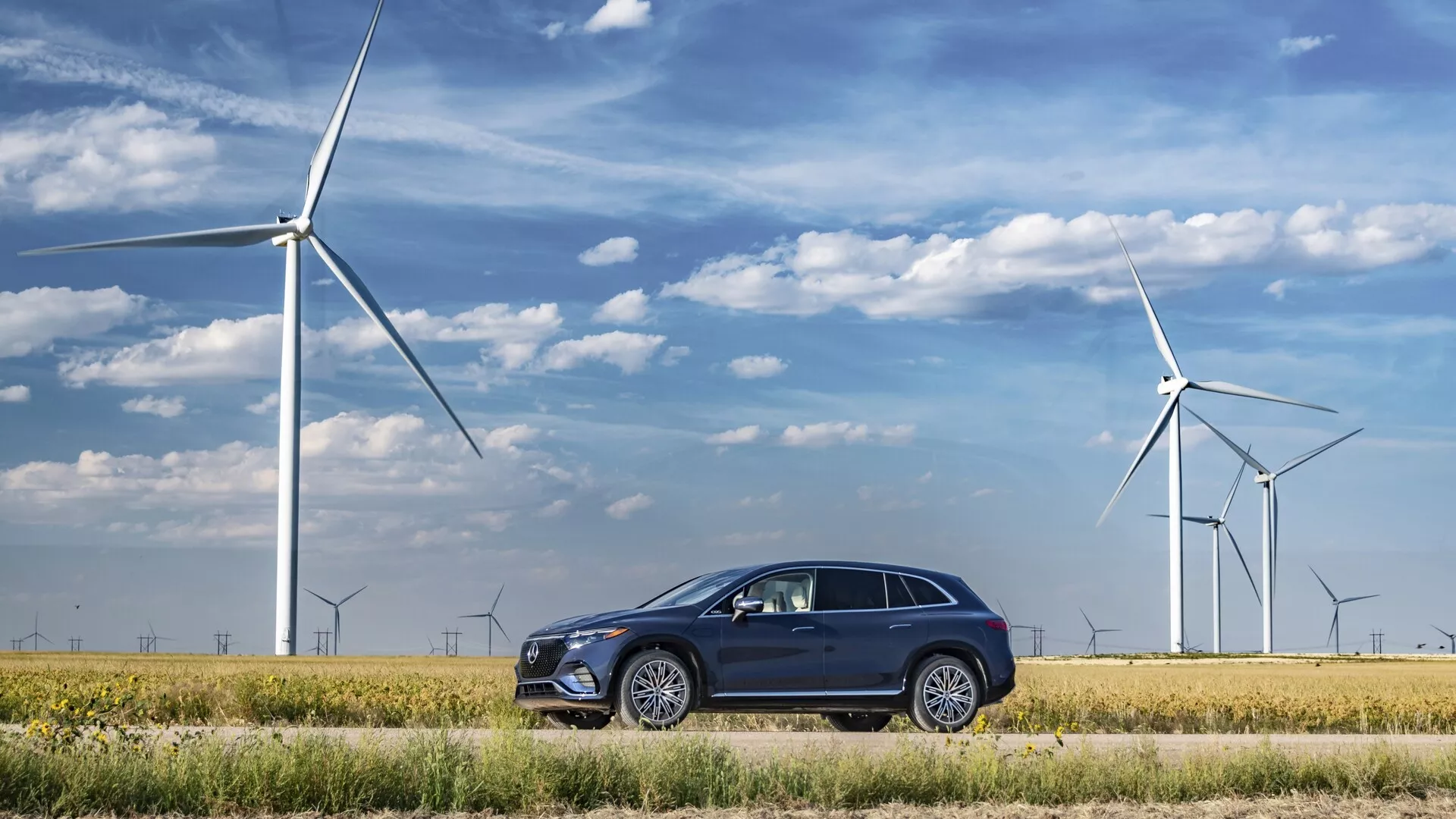 Mercedes EQS-SUV 2022 zaparkovaný v ekologickej krajine s veternými turbínami v pozadí, predstavujúci moderný, elegantný dizajn s tmavým sfarbením, viacramennými kolesami, zapustenými kľučkami dverí, chrómovanými rámčekmi okien, hranatými LED svetlometmi a integrovanými smerovými svetlami v spätných zrkadlách.