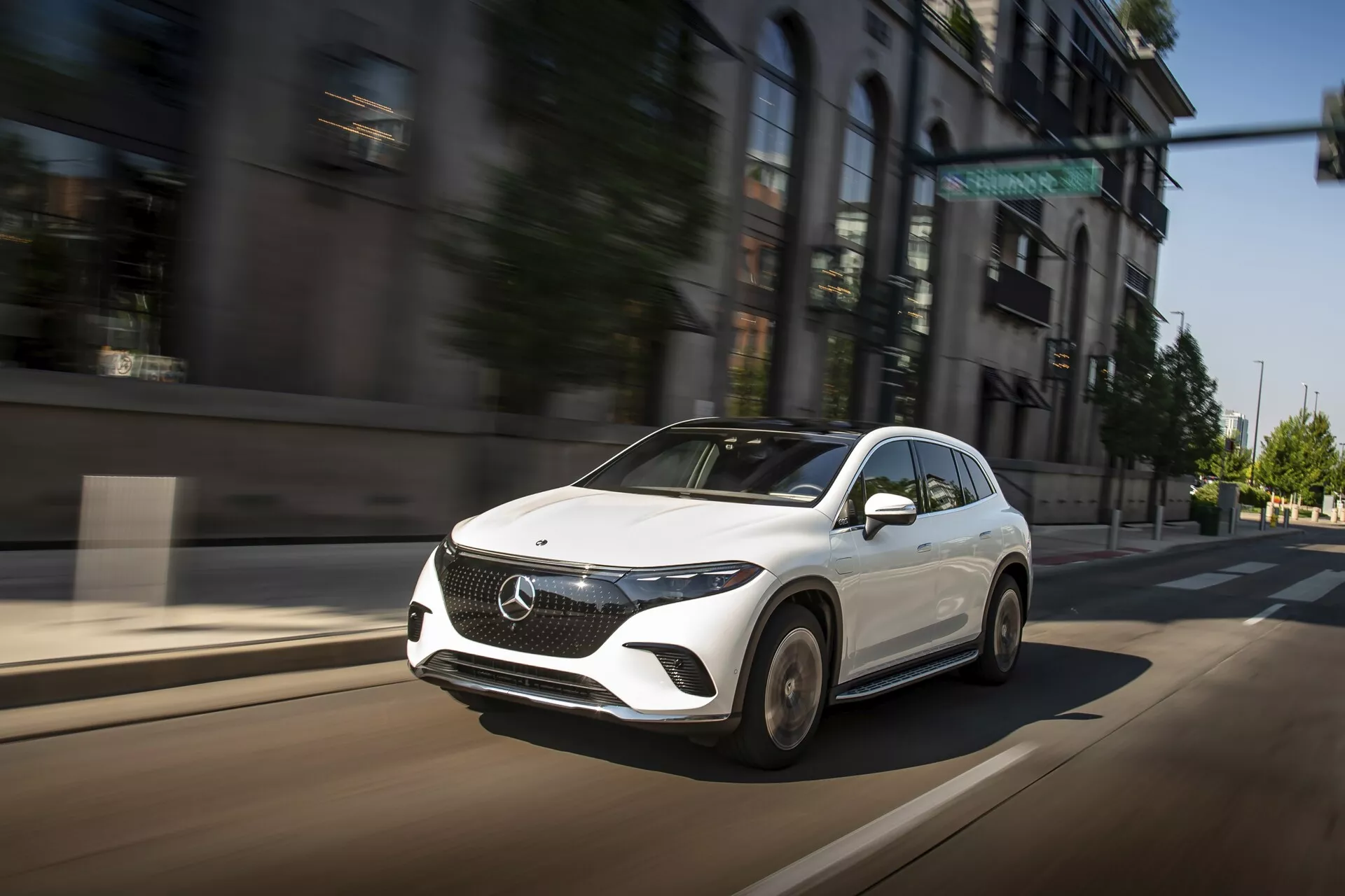 Obrázok zachytáva Mercedes EQS-SUV 2022 v pohybe na mestskej ulici z trojštvrtinového predného uhla, zvýrazňujúci jeho prednú masku, vyklenutú kapotu, elegantné uhlové LED svetlomety a časť dizajnu kolies. Biela farba vozidla kontrastuje s rozmazaným mestským pozadím a zdôrazňuje jeho rýchlosť a dynamickú prítomnosť. Veľký znak na prednej maske, aerodynamicky navrhnuté spätné zrkadlo a pokročilá konfigurácia svetlometov naznačujú jeho moderné, luxusné a technologicky vyspelé vlastnosti.