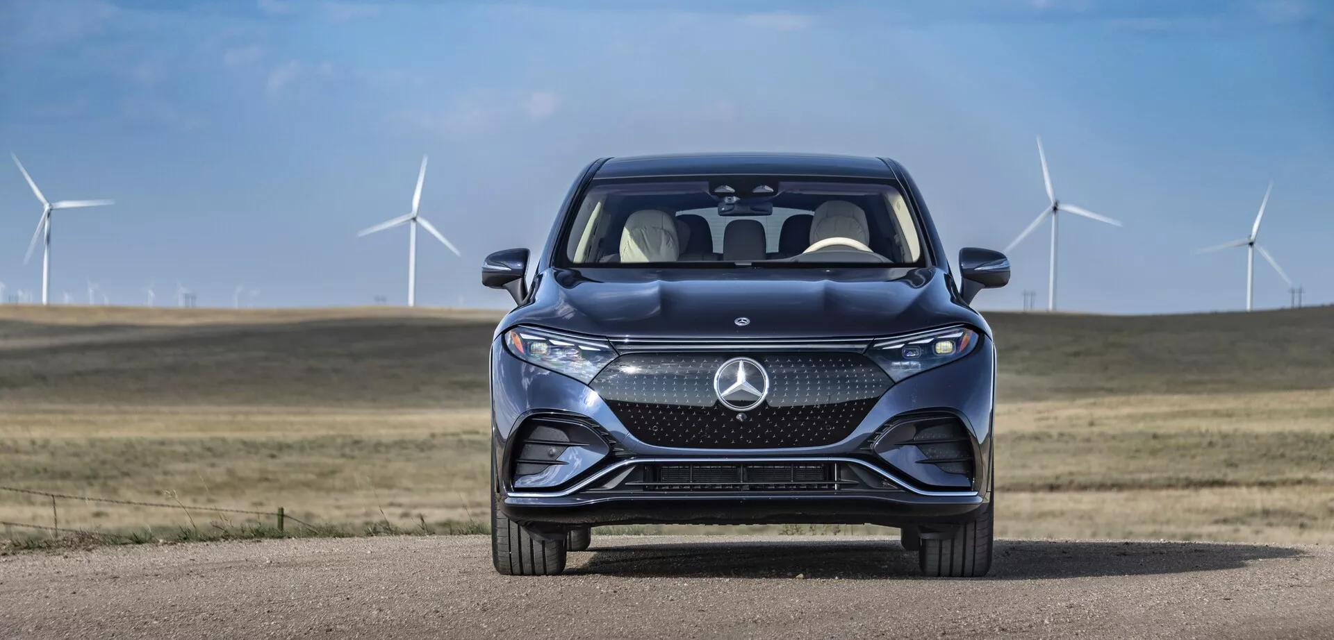 Predný pohľad na Mercedes EQS-SUV 2022 proti pozadiu jasného neba a veterných turbín. Vozidlo má výraznú masku chladiča s veľkým štýlovým logom a zložitým vzorom mriežky, ktorý pravdepodobne obsahuje senzory pre vodičské asistenčné systémy. Sú viditeľné štíhle horizontálne LED svetlá so zabudovanými dennými svetlami. Športový predný nárazník zahrňuje veľké prívody vzduchu a kontúry kapoty sa sklápajú dopredu pre lepšiu aerodynamiku. Čiastočný pohľad na prednú štvrtinu odhaľuje časť blatníkov, čo naznačuje prémiový dizajn. Odraz predného skla a čiastočne viditeľné spätné zrkadlo na strane spolujazdca naznačuje pokročilé funkcie, ako napríklad smerové svetlá a monitorovanie slepého uhla. Pokročilé vodičské asistenčné systémy a náznaky trvalo udržateľného prostredia sú naznačené okolitými veternými turbínami.