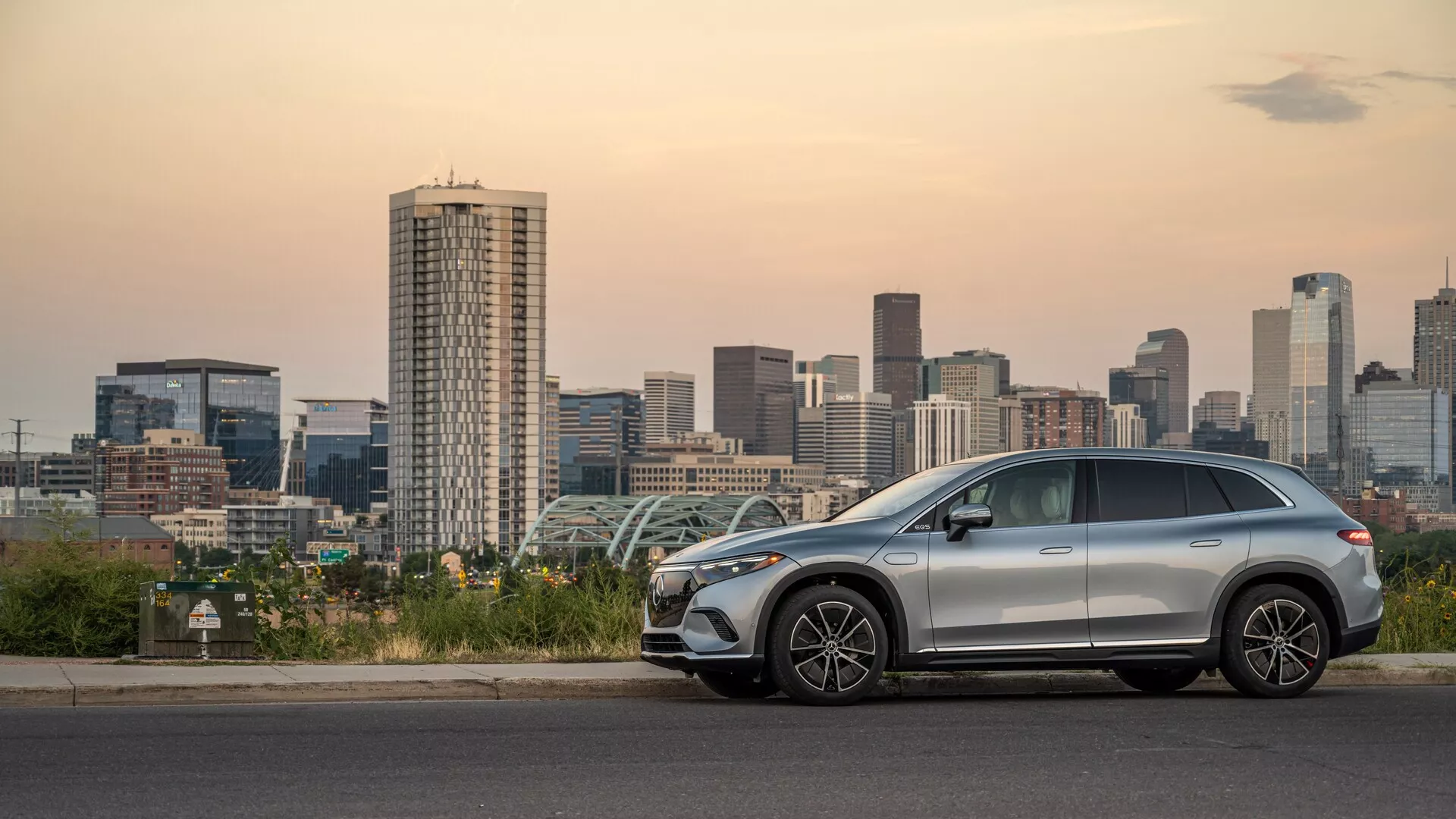 Bočný pohľad na moderné stredne veľké SUV Mercedes EQS 2022 zaparkované pri mestskom panoráme za súmraku, ktorý zobrazuje elegantný aerodynamický dizajn, hliníkové kolesá, strešné nosiče, možné strešné okno a pokročilé funkcie, ako sú integrované smerovky a bezkľúčový vstup.