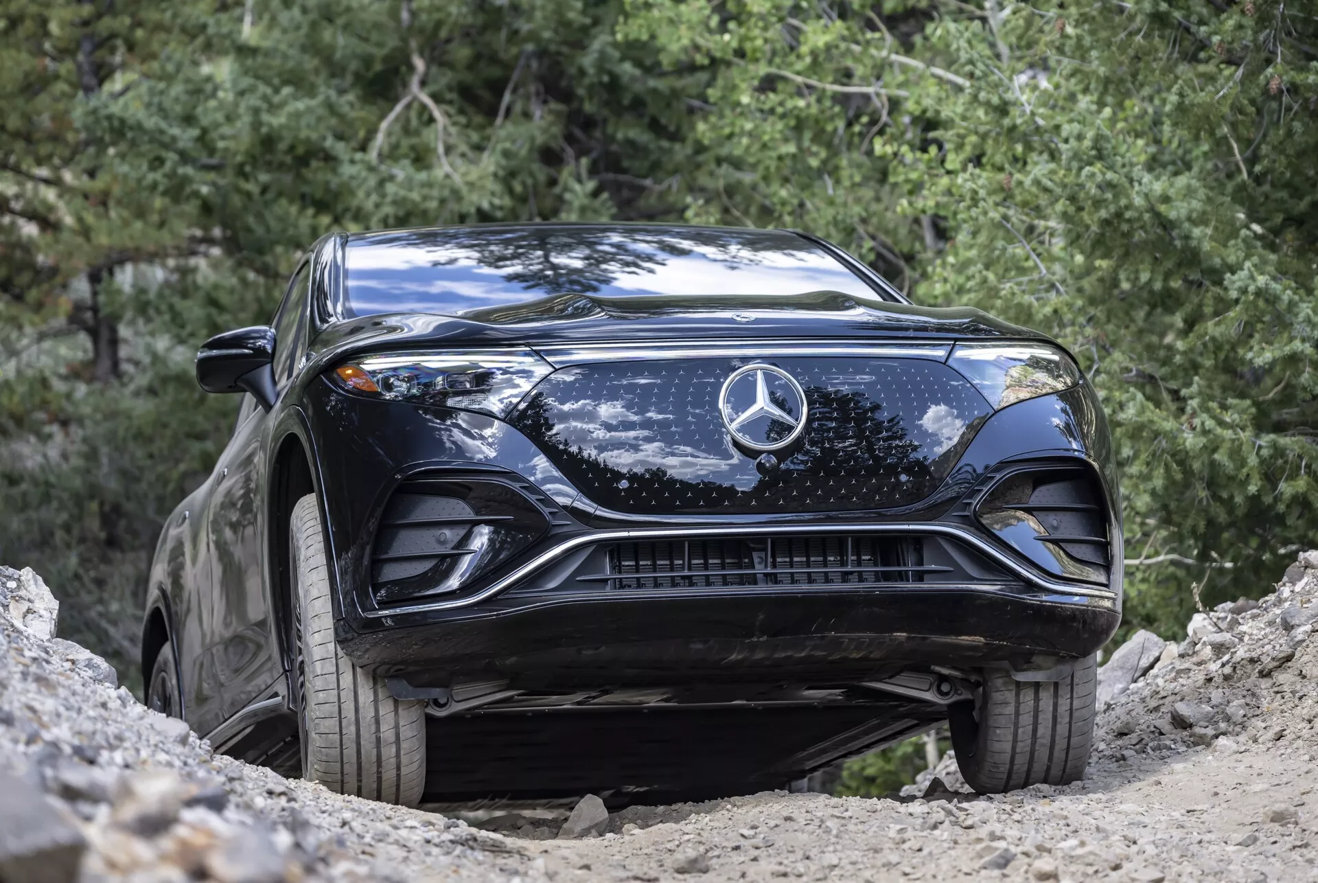 Záber z nízkeho uhla na čierny Mercedes-Benz EQS SUV 2022, zvýrazňujúci jeho tvarovanú kapotu, veľké centrálne hviezdne logo, prepracovanú mriežku, elegantné LED svetlomety a spodné prívody vzduchu. Vozidlo je zachytené v prírodnom vonkajšom prostredí, umiestnené na svahu, čo zdôrazňuje jeho vyššiu svetlú výšku a potenciál pre jazdu v teréne. Vozidlo je čisté s niekoľkými škvrnami od špiny, čo podčiarkuje jeho ľahké terénne využitie a pokročilý automobilový dizajn.