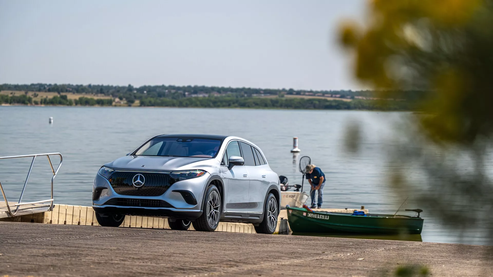 Obrázok zobrazuje luxusný Mercedes-Benz EQS SUV 2022 zaparkovaný pri jazere alebo na riečnom móle. Vozidlo je mierne naklonené, čo umožňuje pohľad z troch štvrtín prednej časti, ktorý zvýrazňuje charakteristickú mriežku, detailné LED svetlá, predný nárazník, čelné sklo a časť zadnej časti. Prostredie je pokojné s vodnou plochou a ďalšími plavidlami v pozadí, čo naznačuje rekreačný kontext. Scéna je dobre osvetlená, čo zdôrazňuje elegantné línie a tvary vozidla, s rozmazanými listami vytvárajúcimi prirodzený rám.