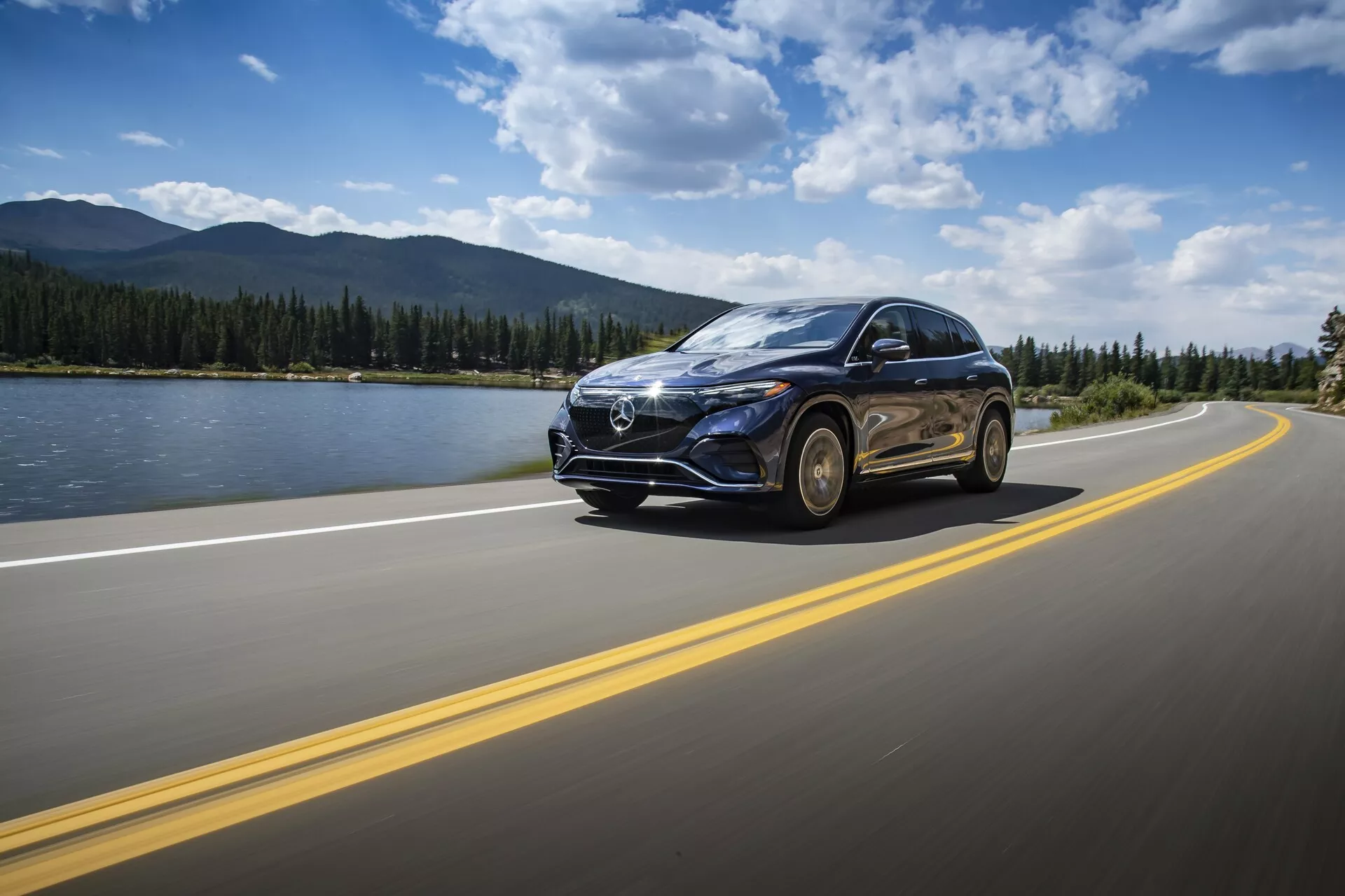Mercedes EQS SUV 2022 jazdí po asfaltovej ceste s dvojitými žltými čiarami pod čiastočne zamračenou oblohou počas dňa. Vozidlo je zachytené z nízkeho predného trojštvrťového uhla, zdôrazňujúc prominentnú prednú mriežku s veľkým symbolom, LED svetlami, elegantné línie kapoty, zliatinové kolesá a bočné zrkadlo s možnými integrovanými smerovkami. Pozadie obsahuje pokojný krajinný výjev s vodným útvarom, horami a čistým horizontom, zvýrazňujúc pocit cestovania a dobrodružstva. Pohyb auta je zdôraznený miernym pohybovým rozmazaním.
