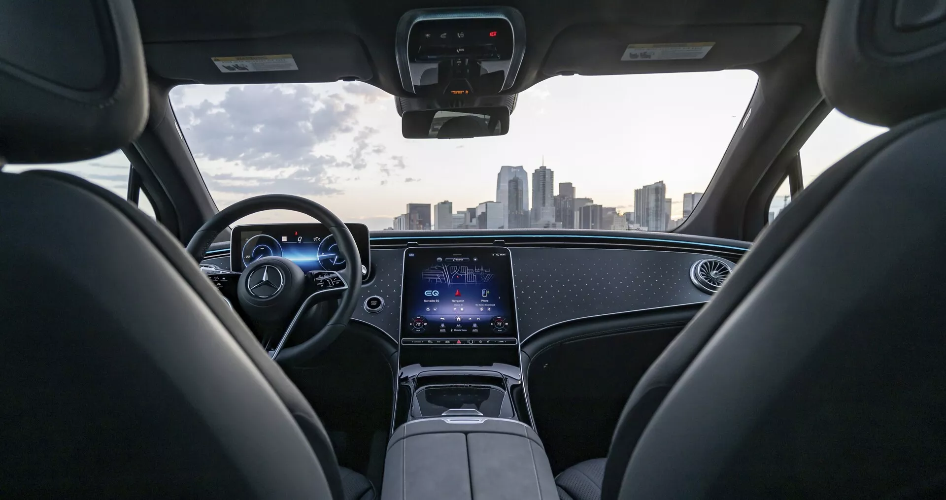 Pohľad na interiér vozidla Mercedes EQS-SUV 2022 z perspektívy zadného sedadla, ukazujúci modernú prístrojovú dosku s ambientným osvetlením, digitálnym prístrojovým panelom a koženým volantom s integrovanými ovládacími prvkami. Stredovej konzole dominuje veľká dotyková obrazovka pre infotainment a pripojovacie funkcie. Čelné sklo poskytuje jasný a neobmedzený výhľad na mestskú panorámu, zdôrazňujúc pokročilú technológiu a luxusný dizajn vozidla.