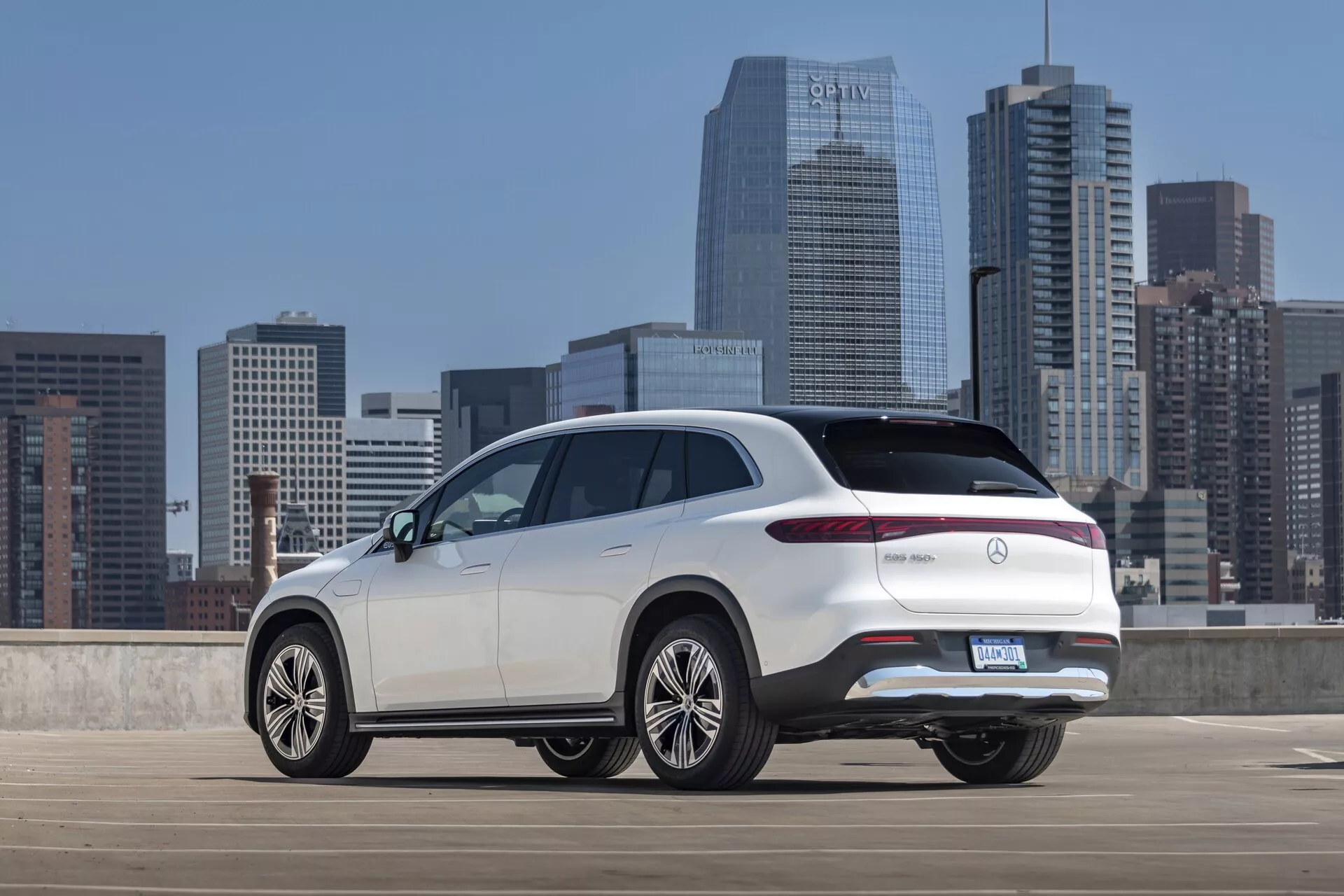 Trojštvrťový zadný pohľad na biely Mercedes EQS-SUV 2022 z mierne zvýšeného uhla, ktorý zobrazuje hornú časť zadnej časti, bočný profil a výrazné dizajnové prvky ako elegantné zadné LED svetlá, zadné dvere, zadný nárazník a veľké viacramenné zliatinové kolesá. SUV je zaparkované v otvorenej mestskej parkovacej štruktúre s mestskou panorámou v pozadí, vyzdvihujúc jeho moderný, luxusný dizajn s potenciálnymi pokročilými vlastnosťami typickými pre luxusné modely.