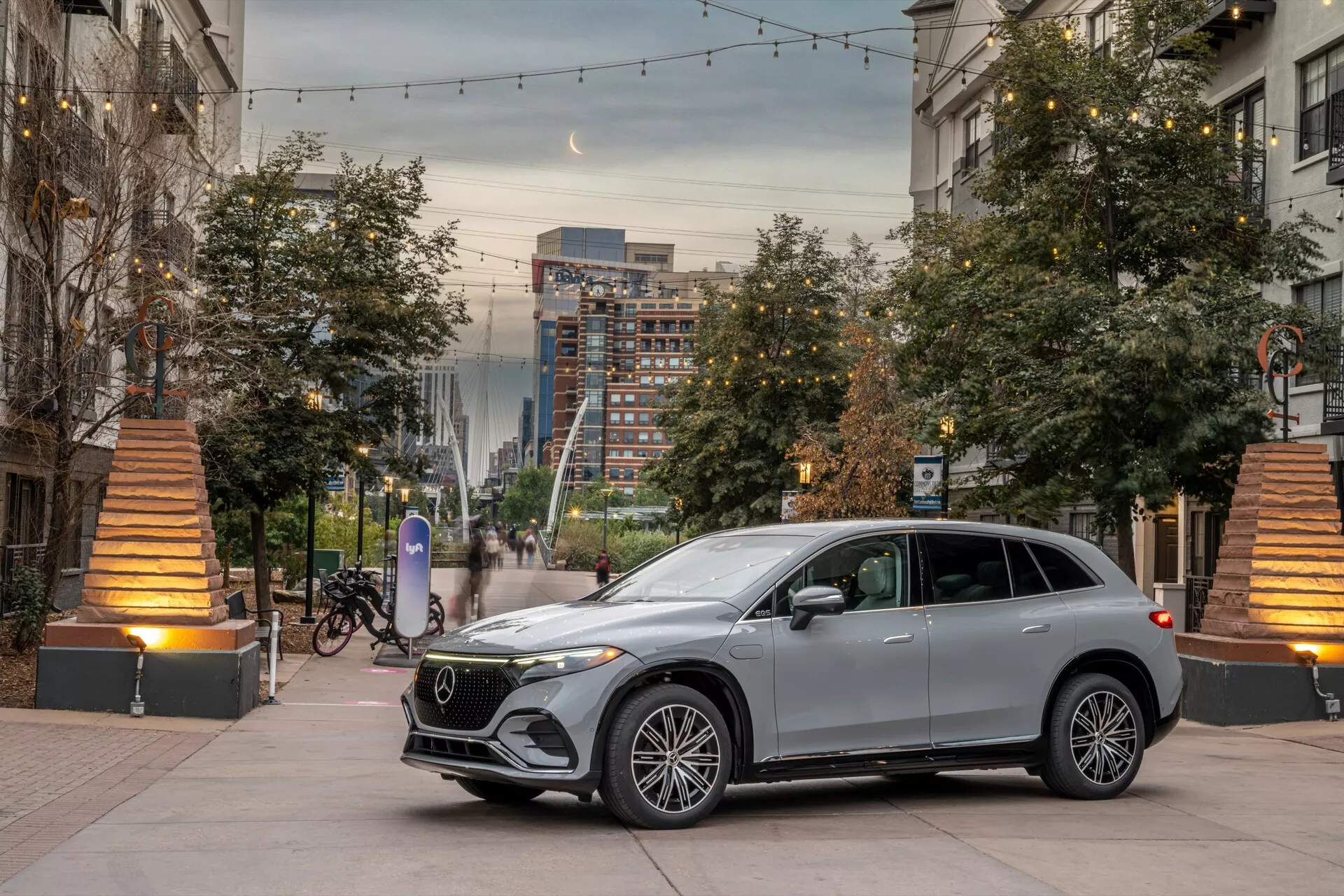 Moderné SUV v mestskom prostredí počas svitania alebo súmraku, snímka z nízkeho uhla prednej štvrtiny zvýrazňujúca prednú mriežku, svetlomety, kapotu a časť vodičovej strany. Centrálna kompozícia s kvetináčmi a teplými pouličnými svetlami pridáva hĺbku. Vynikajúce luxusné prvky zahŕňajú viacramenné zliatinové kolesá, LED svetlomety, mriežku s veľkým emblémom, integrované zrkadlové smerovky, elegantné dizajnové línie a metalický lak.