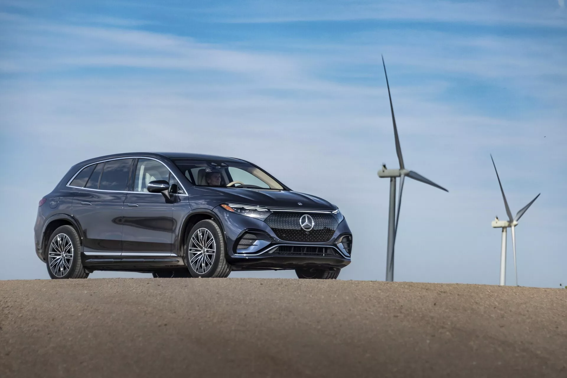 Trojtretinový predný pohľad na šedý Mercedes EQS-SUV 2022 postavený na malom kopci, ktorý ukazuje moderný dizajn s elegantnými líniami karosérie, výraznými tvarmi svetlometov, výraznou prednou maskou s emblémom loga, nehybnými kolesami a kolieskami z ľahkých zliatin. Veterné turbíny v pozadí naznačujú veternú farmu alebo vidiecku oblasť, čo možno naznačuje ekologické vlastnosti vozidla.