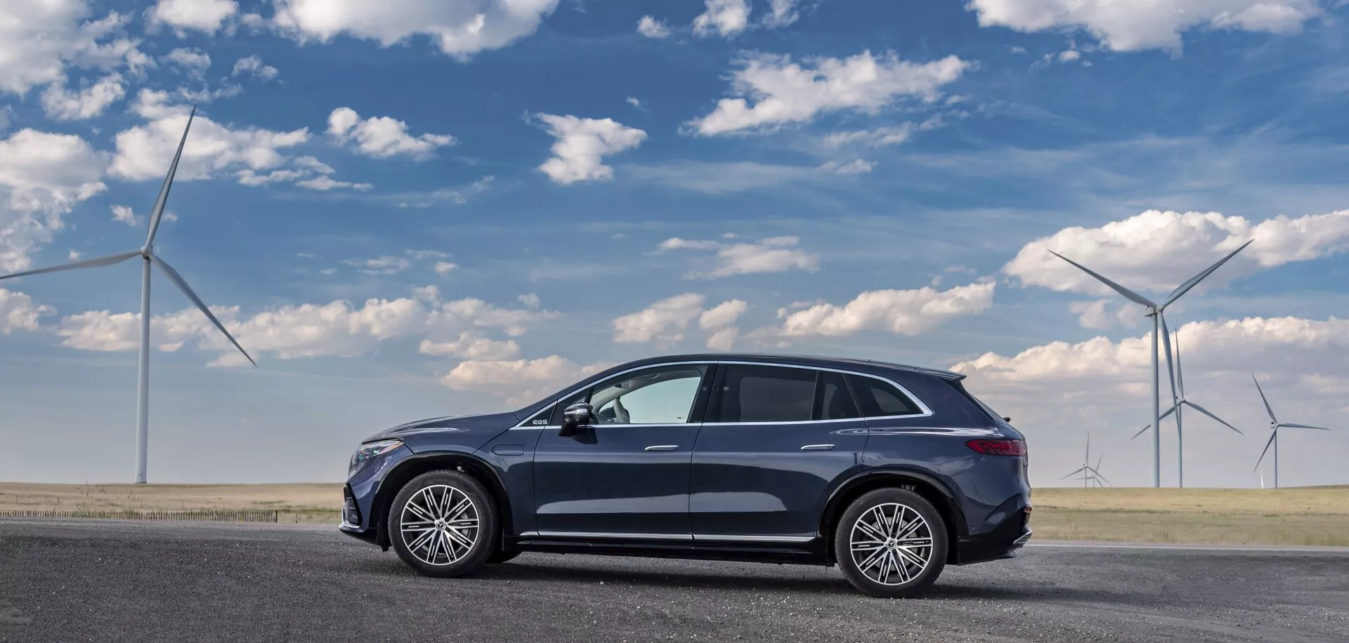 Pohľad zboku na tmavé Mercedes EQS-SUV 2022 zaparkované na rovnom povrchu cesty, zachytené z pohľadu spolujazdca. Elegantný, aerodynamický moderný crossover SUV má päť dverí, veľké viacramenné zliatinové kolesá, panoramatické strešné okno, strešnú anténu a spätné zrkadlá s integrovanými smerovkami. Jeho profil naznačuje zvýšený postoj typický pre crossovery. Veterné turbíny v pozadí naznačujú ekologické témy.