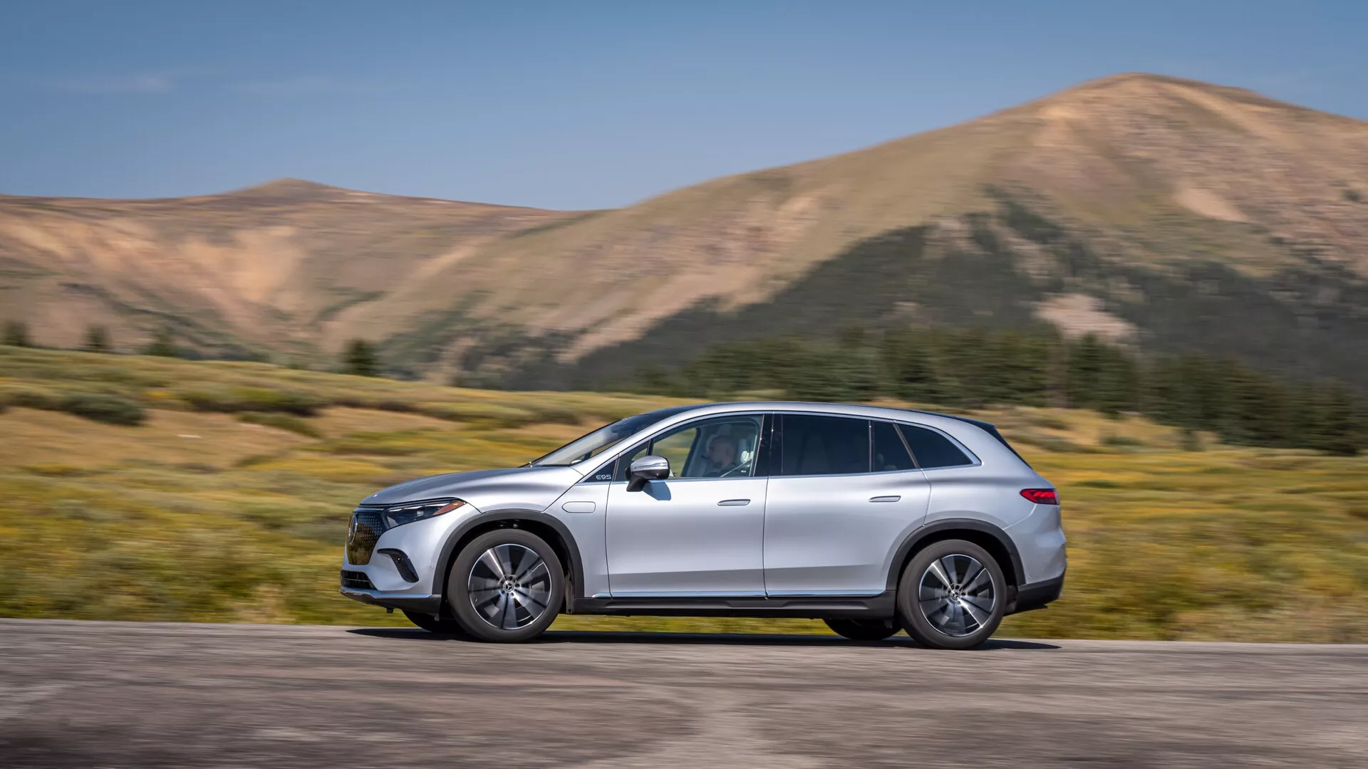 Mercedes EQS-SUV 2022 zachytený v bočnom pohľade, umiestnený v centrálnej časti rámu s pohybom zrejmým z mierne rozmazaného pozadia. Plnohodnotné SUV so štýlovým, zakriveným dizajnom, aerodynamickými bočnými zrkadlami, liatinovými kolesami, strešnými nosičmi, chrómovými prvkami a metalickým lakom. Viditeľní sú pokročilé technologické prvky vrátane LED denných svetiel a zapustených kľučiek dverí, naznačujúcich bezkľúčový vstup. Horská krajina umocňuje dynamický a dobrodružný vzhľad, ideálny pre automobilové inzeráty a životný štýl.