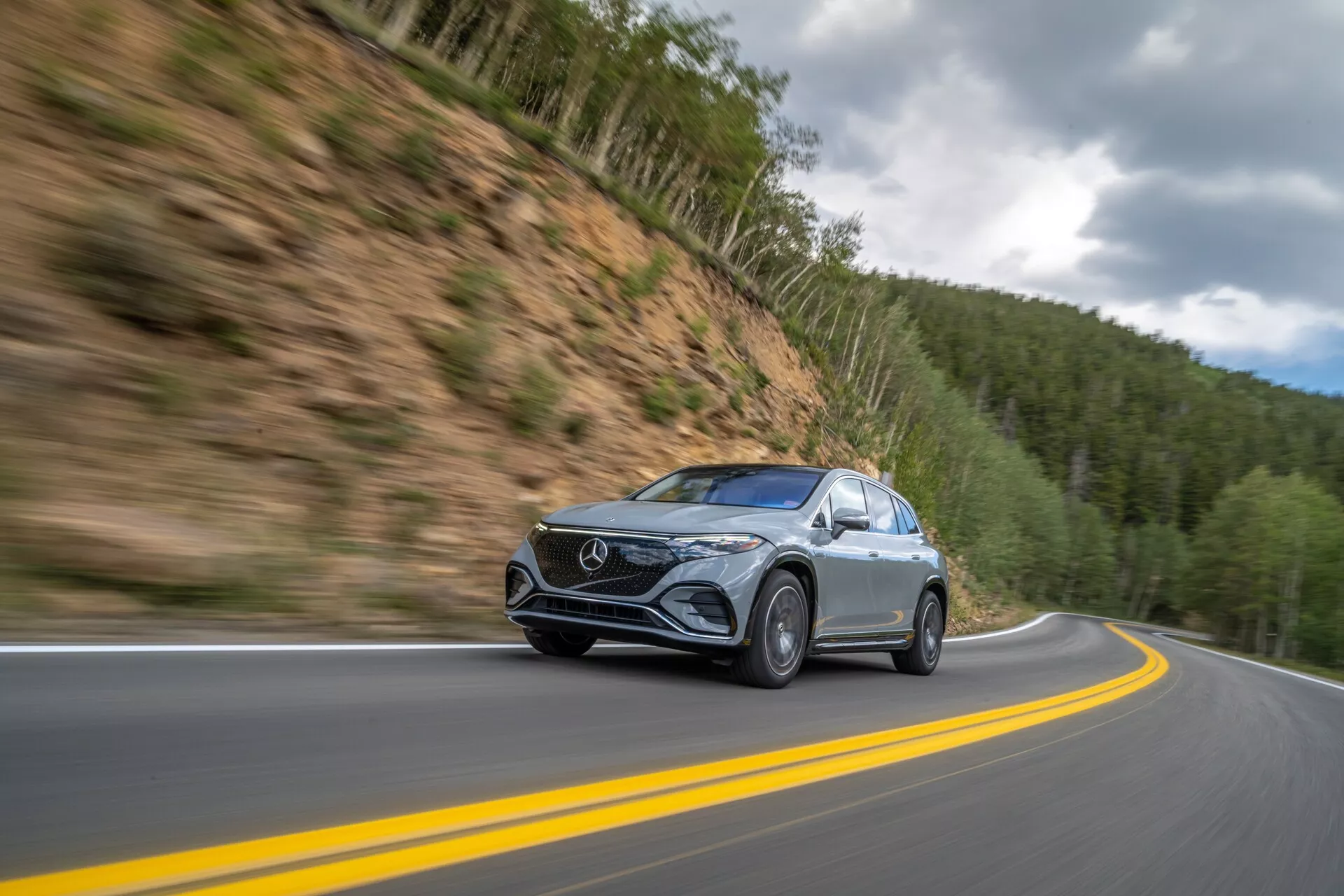Dynamický záber Mercedes EQS-SUV 2022 v pohybe na horskej ceste, zachytený z nízkeho, mierne predného uhla. Auto, umiestnené vľavo od stredu, sa pohybuje po kľukatej dvojprúdovej ceste s rozmazaným pozadím, čo naznačuje vysokú rýchlosť. So štýlovými LED svetlometmi, výraznou prednou maskou a kovovým lakom vozidlo vyniká na pozadí lesnatého hôr pod zamračenou oblohou. Detaily, ako sú rozmazané kolesá v pohybe a jasné bočné zrkadlá s možnými integrovanými smerovými svetlami, zvyšujú technologickú príťažlivosť.