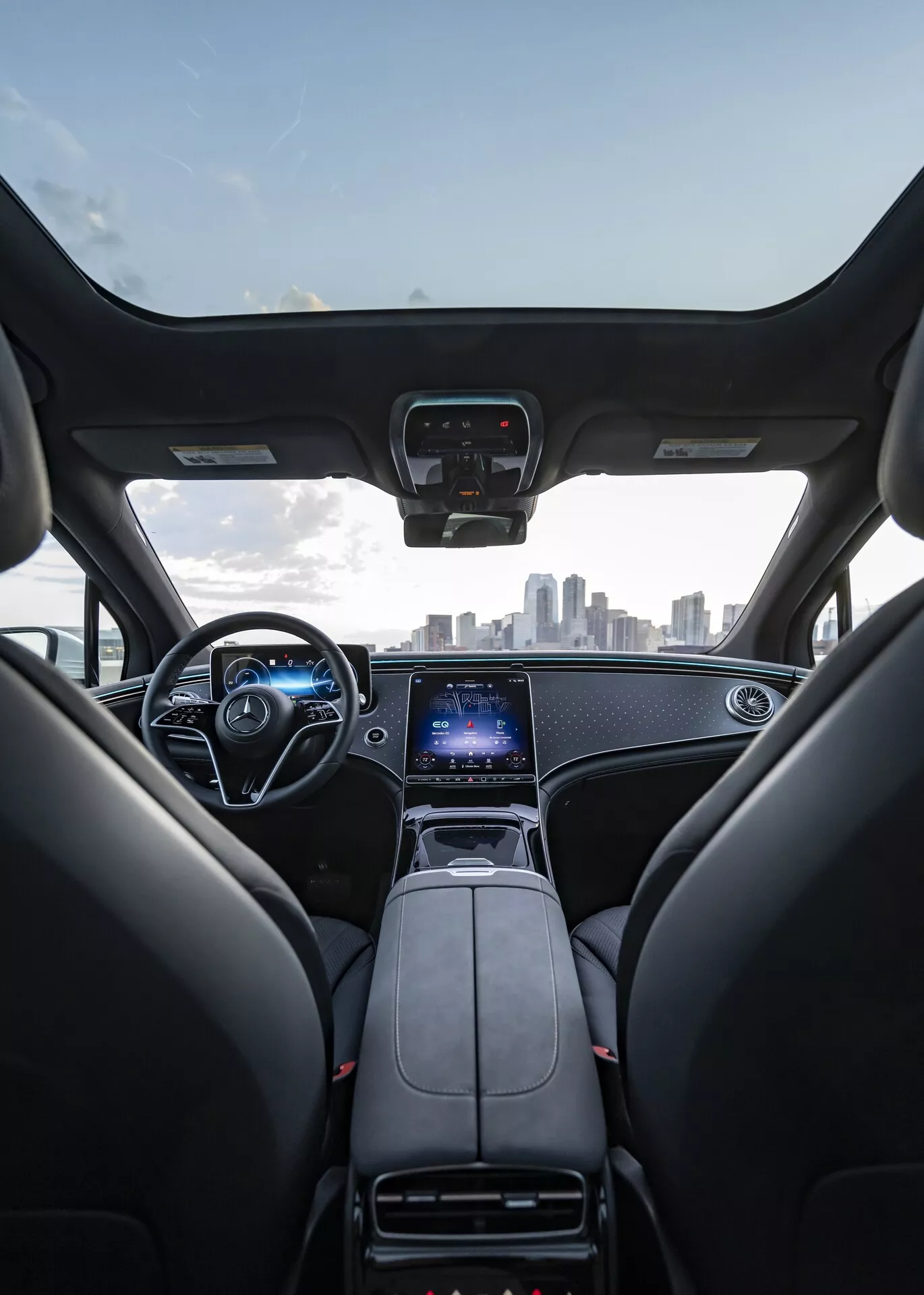 Pohľad na interiér Mercedes EQS SUV 2022 z perspektívy zadného sedadla, ukazujúci predné sedadlá, palubnú dosku, volant a čelné sklo. Predné sedadlá sú z čiernej kože s opierkou hlavy, palubná doska má elegantný dizajn s kovovými akcentmi. Stredová konzola obsahuje radiacu páku, držiaky na nápoje a veľký vertikálne orientovaný dotykový displej pre infotainment, GPS a nastavenia vozidla. Volant zahŕňa viacero ovládacích tlačidiel a za ním je viditeľný digitálny prístrojový panel. Obrázok tiež zachytáva mestskú panorámu cez čelné sklo, čo naznačuje prémiový, moderný interiér s pokročilou technológiou.