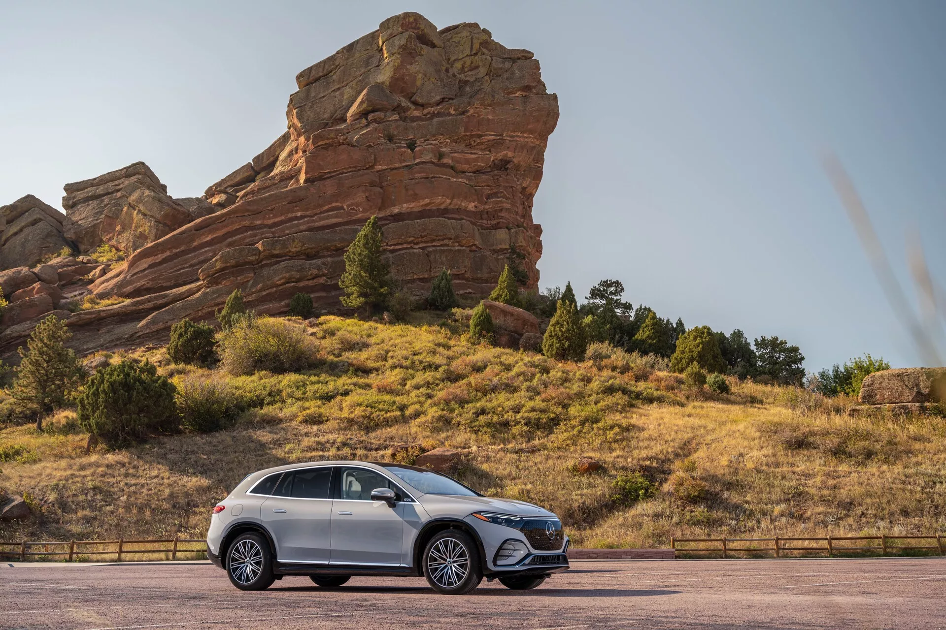 Bočný profilový pohľad na Mercedes EQS SUV 2022 zaparkovaný na asfaltovej ceste s veľkými skalnými útvarmi v pozadí, zdôrazňujúci jeho vyššiu svetlú výšku, disky z ľahkej zliatiny, elegantné dizajnové línie, zatmavené okná a lesklý lak.
