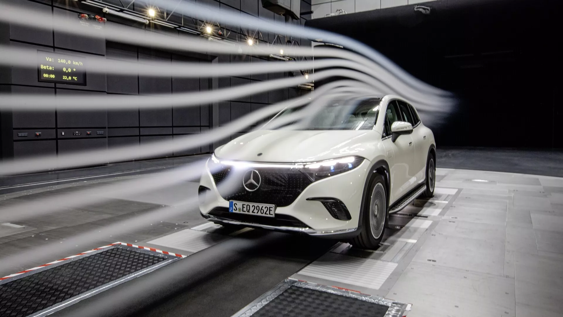 Obrázok bieleho vozidla Mercedes EQS-SUV 2022 počas testovania v profesionálnom zariadení. Auto, umiestnené uprostred snímky pod miernym uhlom, ukazuje svoju prednú mriežku, LED svetlomety, kapotu, čelné sklo a časť strany vodiča. Čiary rozmazania pohybu naznačujú aerodynamické testovanie. Futuristický dizajn, veľké logo na mriežke a vnútorné testovacie prostredie.