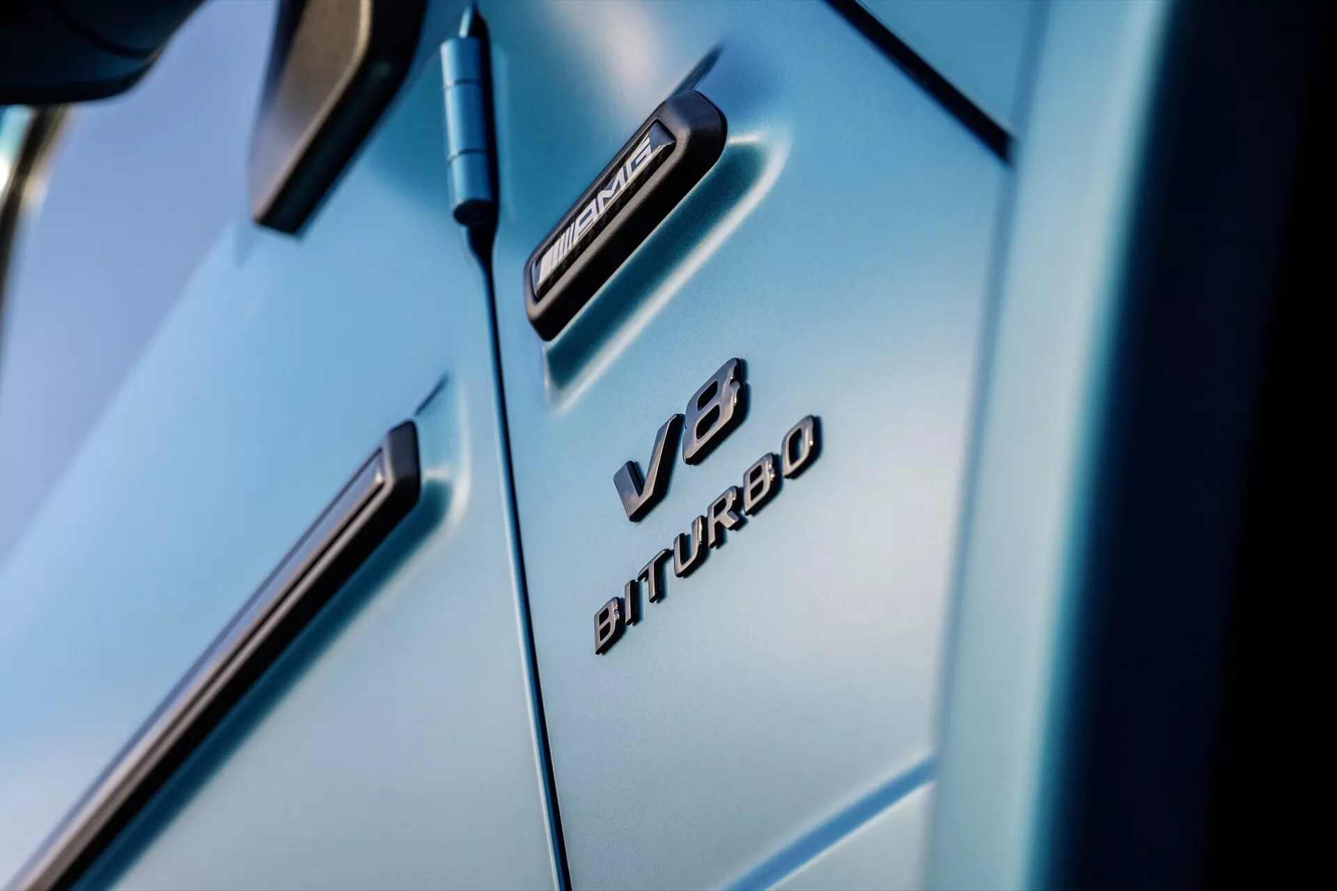 Detailný pohľad na predný blatník Mercedes-Benz G-Klasse 2024 s odznakmi V8 BITURBO a 4MATIC, ktorý zdôrazňuje luxusné dizajnové línie a lesklý svetlomodrý povrch