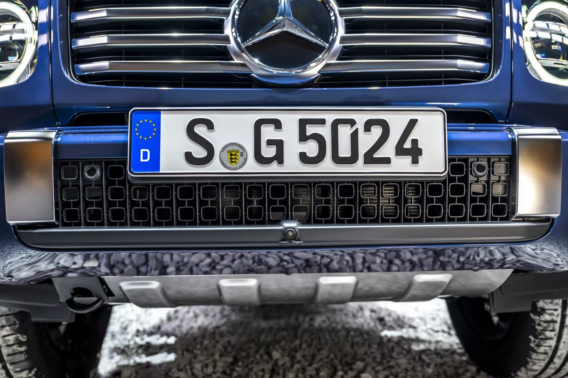 Detailný pohľad na prednú časť Mercedesu-Benz triedy G rok 2024 s mriežkou zdobenou čiernymi štvorcami s chrómovými hranami, európskou evidenčnou značkou, robustným predným nárazníkom s kovovou ochrannou platňou a centrálnym odznakom Mercedes-Benz s potenciálnou snímacou technológiou, natretý tmavomodrou farbou, naznačujúci luxusné alebo vysokovýkonné SUV v bezchybnom stave.