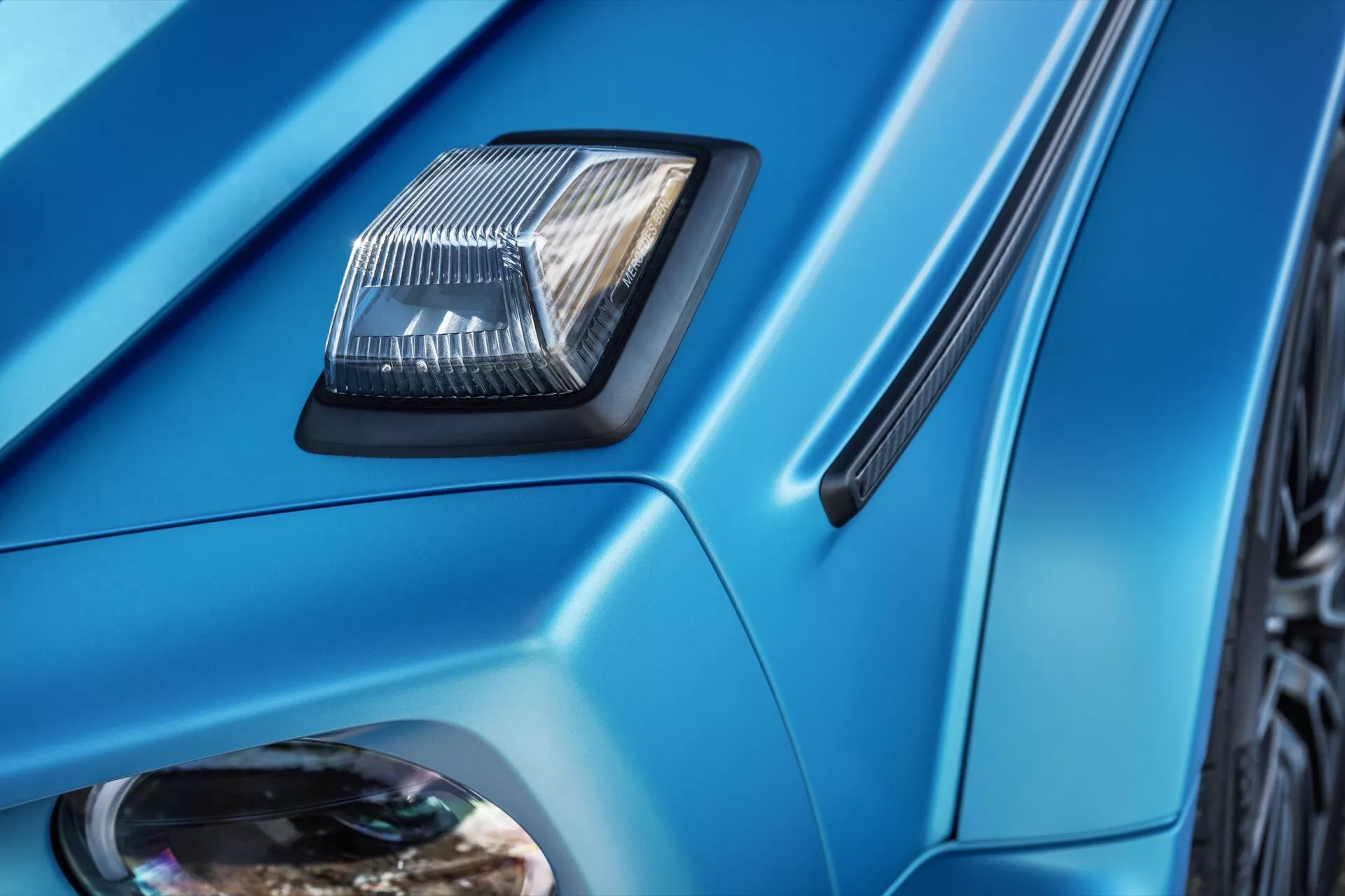 Detailný záber Mercedes-Benz G-Klasse 2024 s otvorom na kapote, ostrými líniami karosérie, metalickou modrou farbou a bočným ventilom pozoruhodné pre designové a technologické prvky