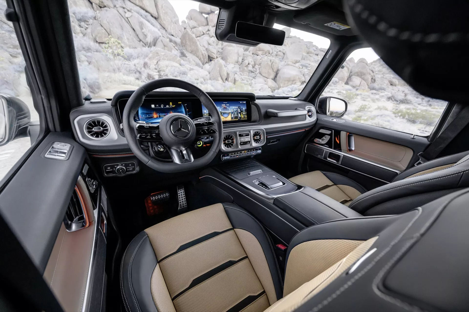 Interiérový pohľad na Mercedes-Benz G-Klasse 2024 (465) zobrazujúci oblasť kokpitu vodiča s multifunkčným volantom, digitálnym prístrojovým panelom, veľkým dotykovým informačno-zábavným systémom, luxusnými sedadlami z dvojfarebnej kože a pokročilými bezpečnostnými funkciami z pohľadu zadného pasažiera