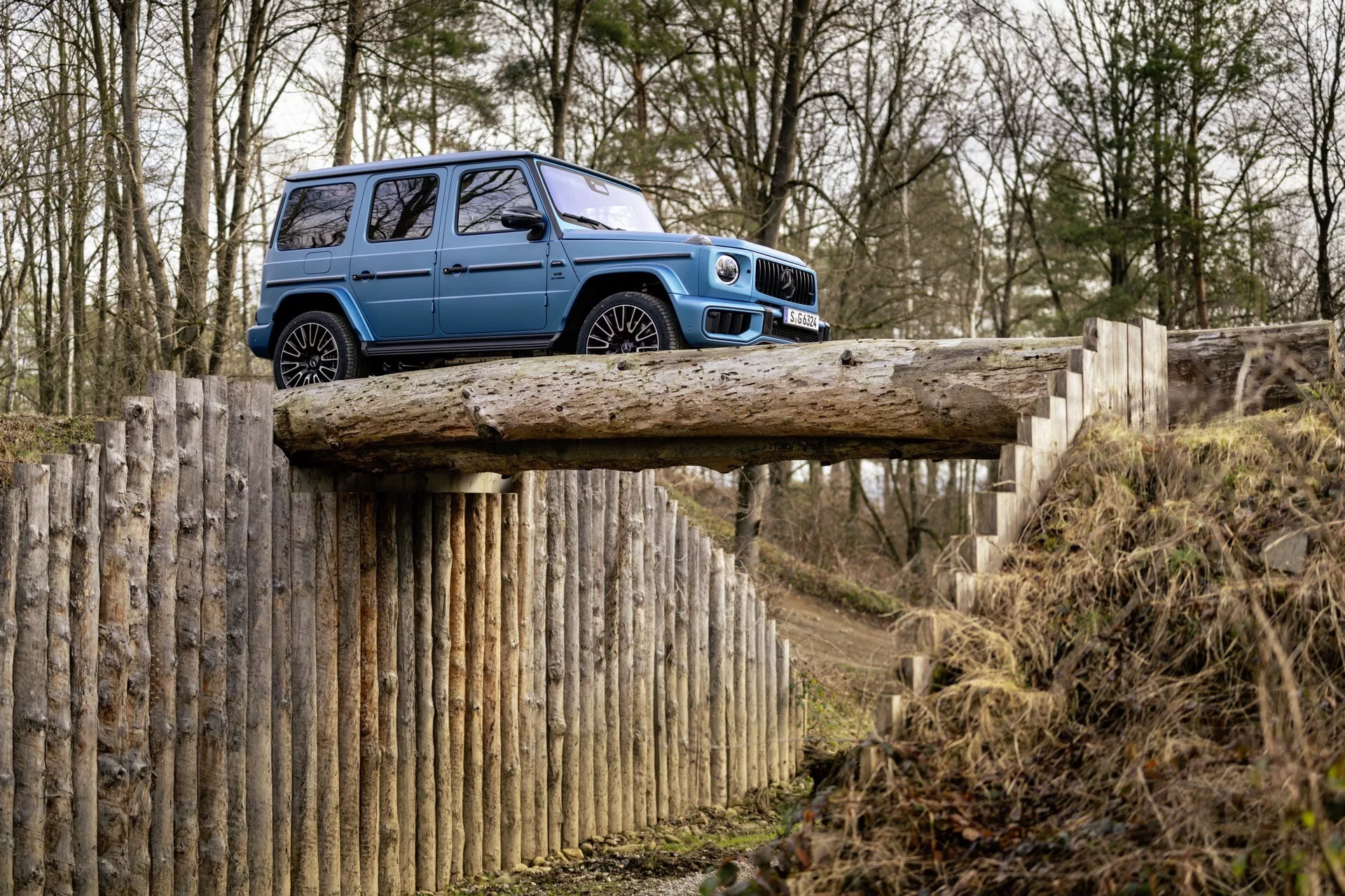 Modré SUV Mercedes-Benz triedy G z roku 2024 na drevenom moste ukazujúce schopnosti v teréne, bočný profil s vysokou svetlou výškou, v lesnom prostredí