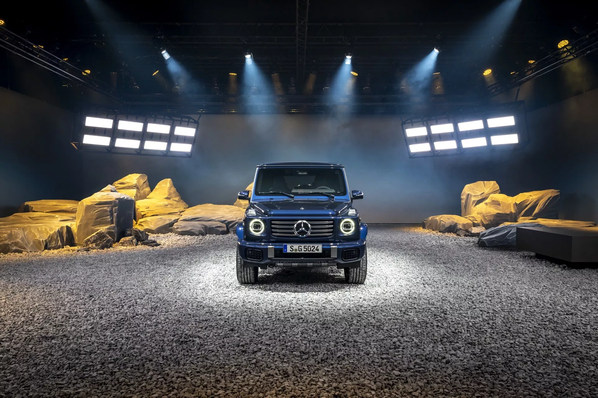 SUV Mercedes-Benz triedy G na rok 2024 na simulovanej vonkajšej scéne so svetelným osvetlením, zobrazujúca prednú mriežku, guľaté svetlomety a robustný dizajn vhodný pre terénne podmienky