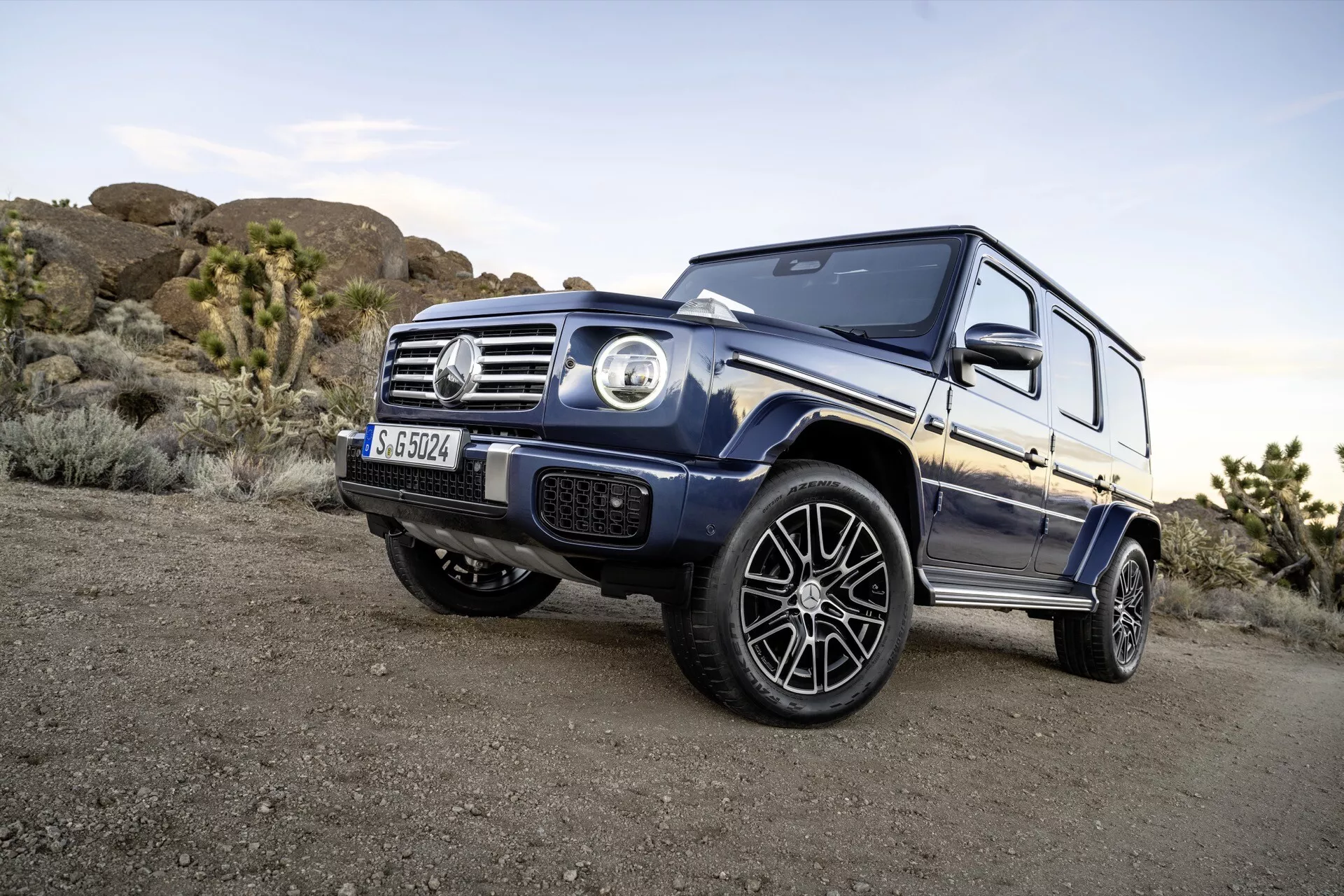 Dynamický uhlový záber na SUV Mercedes-Benz triedy G 2024, ktorý zdôrazňuje jeho dominantný postoj a detailné dizajnové prvky, ako sú okrúhle svetlomety, predná mriežka s emblémom a svalnaté oblúky kolies na pozadí terénu v mäkkom večernom svetle
