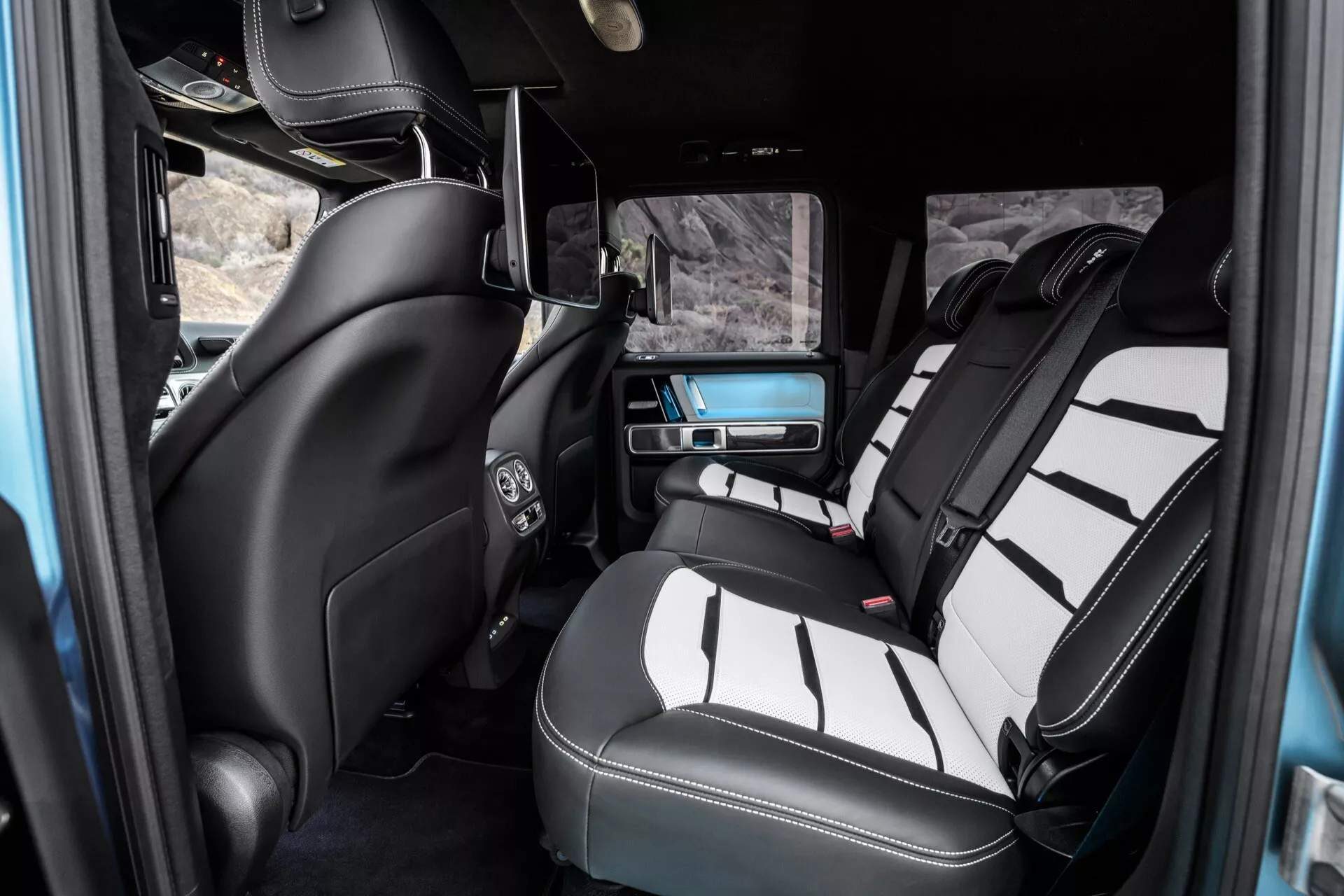 Interiér Mercedes-Benz G-Klasse 2024 s dôrazom na luxusnú zadnú pasažiersku časť, ukazujúc kožené sedadlá v dvoch tónoch, stredovú konzolu, klimatizáciu, zábavný systém a detaily luxusného vybavenia