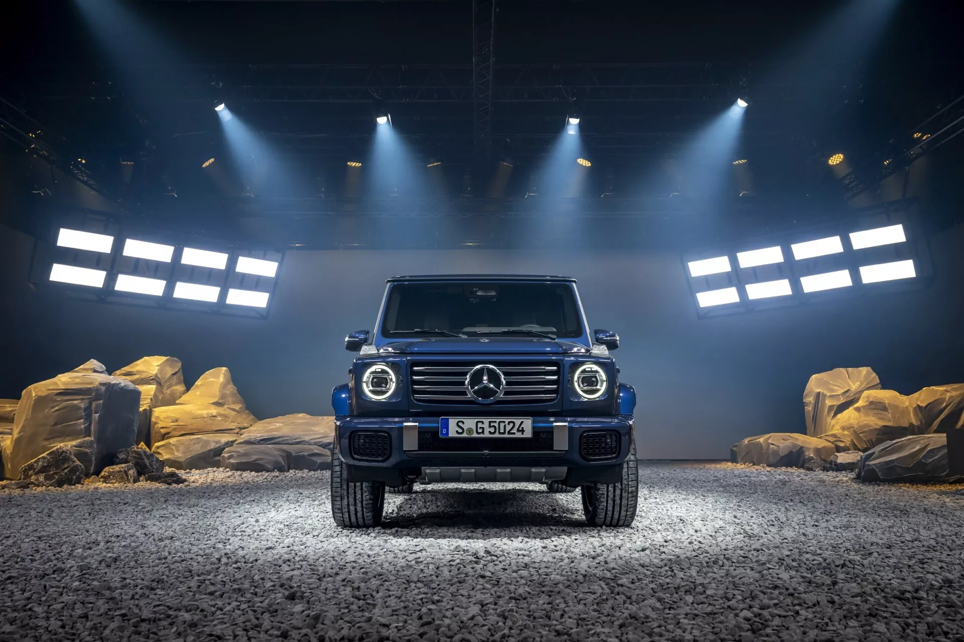 Luxusné terénne SUV Mercedes-Benz triedy G (G-Klasse) 2024 s dramatickým osvetlením, ktoré zdôrazňuje jeho vysokú svetlú výšku, ikonickú mriežku s emblémom, okrúhle LED svetlomety, obdĺžnikové denné svetlá, robustný predný nárazník a svalnatý dizajn kapoty na štrkovom povrchu s dobrodružným štúdiovým pozadím