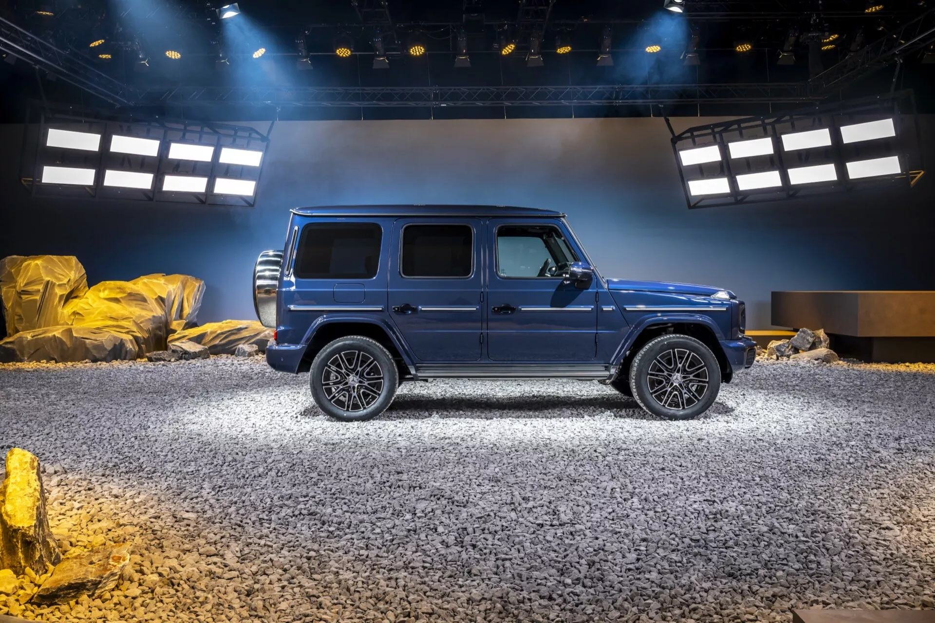 Luxusné SUV Mercedes-Benz triedy G 2024 v metalickej modrej farbe predvádzajúce terénne schopnosti s veľkými zliatinovými kolesami, bočnými stupienkami a strešným oknom, umiestnené v centre simulovaného terénneho výstavného priestoru