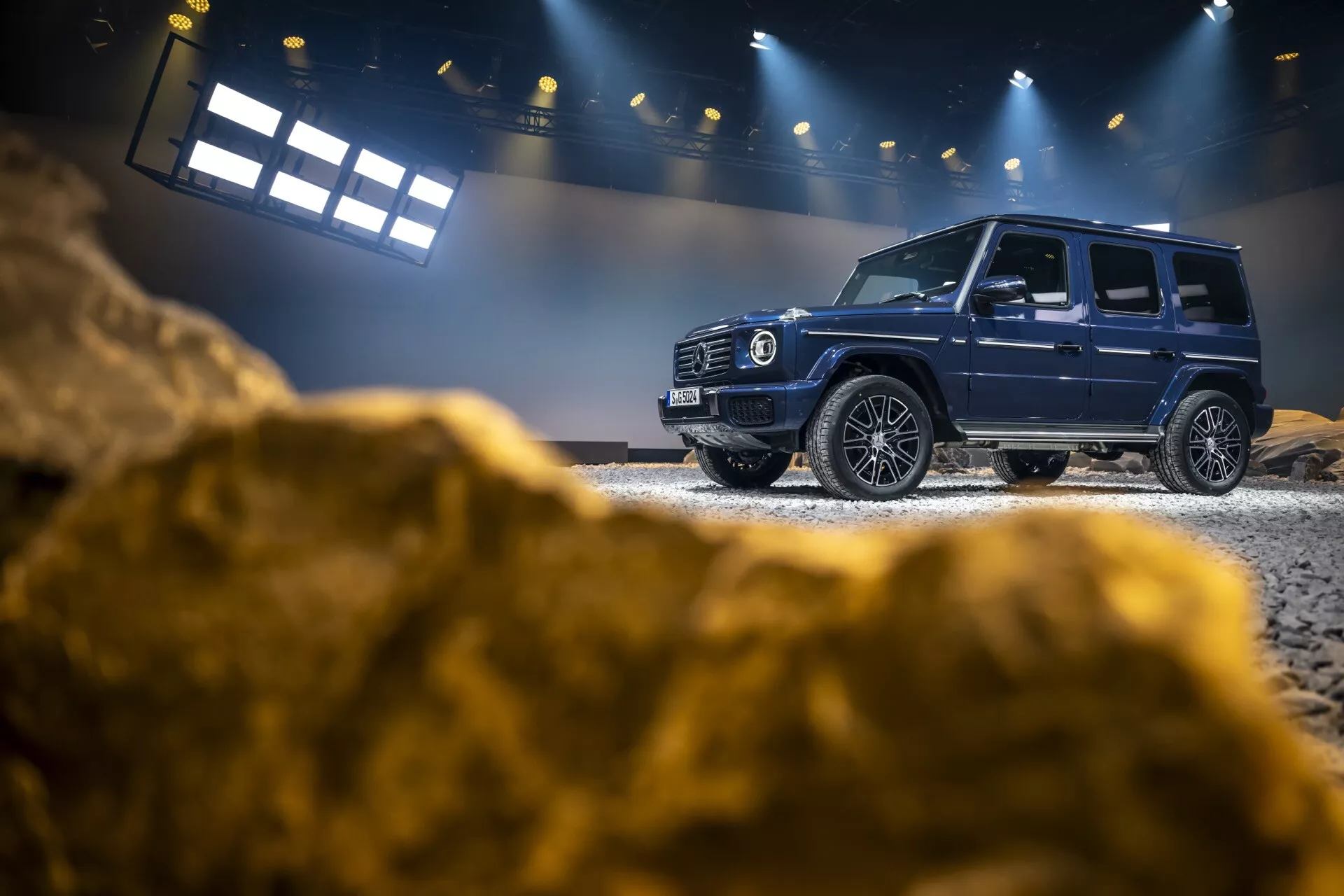 Luxusné SUV Mercedes-Benz triedy G 2024 predstavené v interiéri na simulovanom off-road teréne, zdôrazňujúce bočný profil, prednú mriežku a pokročilé technologické funkcie s kontrolovaným osvetlením a robustným pozadím