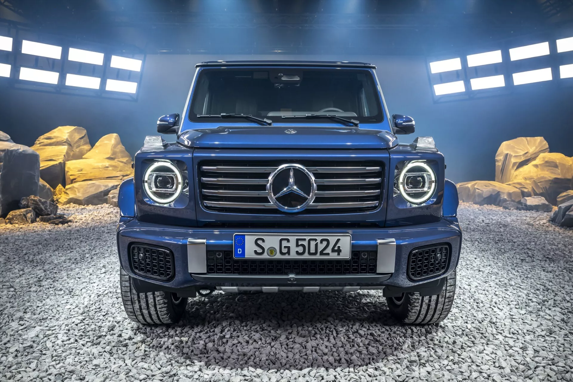 Pohľad zpredu na Mercedes-Benz triedy G 2024 s ikonickou prednou mriežkou, LED svetlometmi, robustným dizajnom kapoty a predným nárazníkom pripraveným na off-road s ochrannou doskou, zachytený v prírodnom vonkajšom prostredí