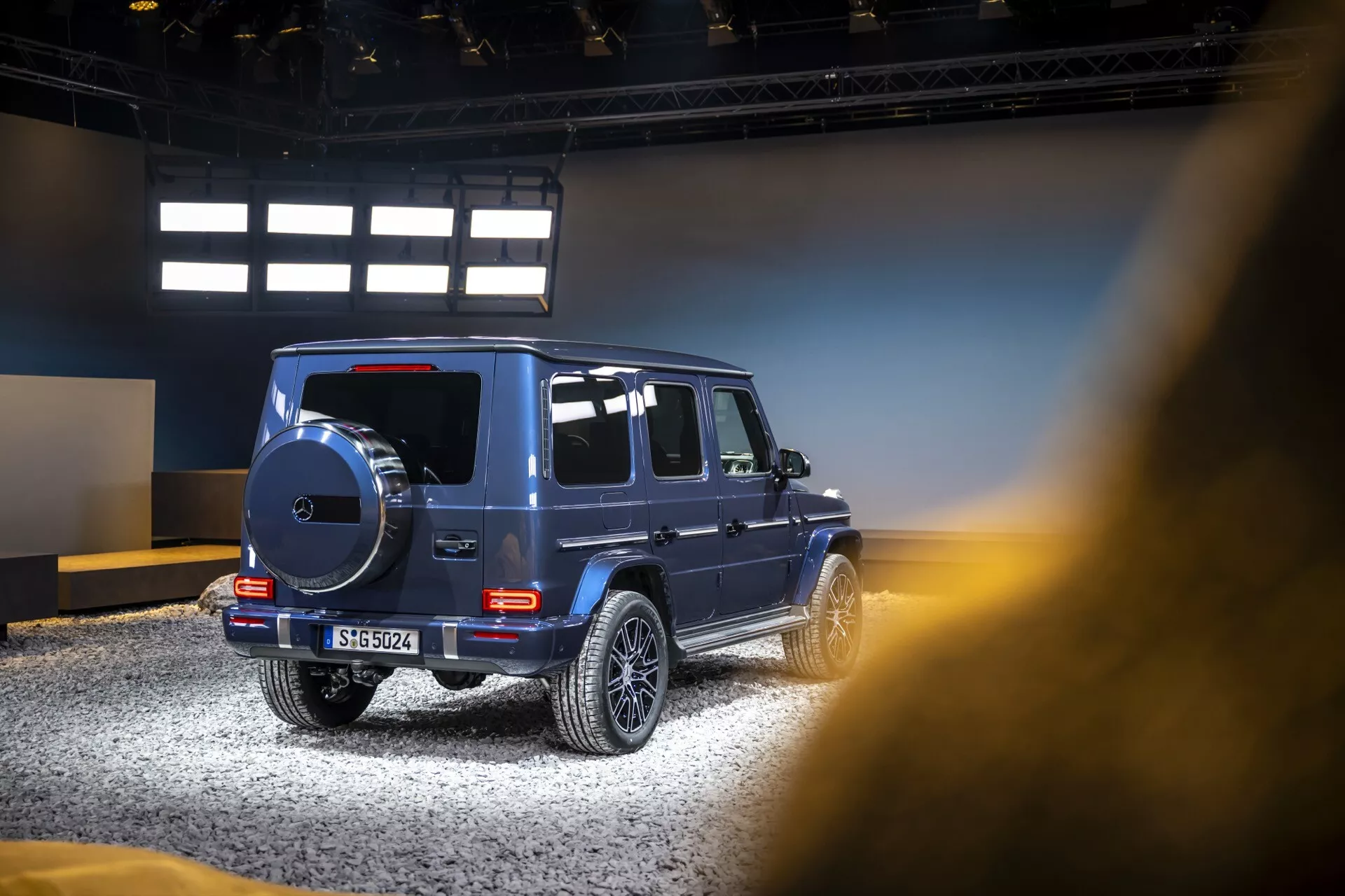 Bočný a čiastočný zadný pohľad na luxusné SUV Mercedes-Benz triedy G 2024, zdôrazňujúci prvky dizajnu pre offroad, zliatinové disky a tónované okná v prostredí vnútornej výstavy